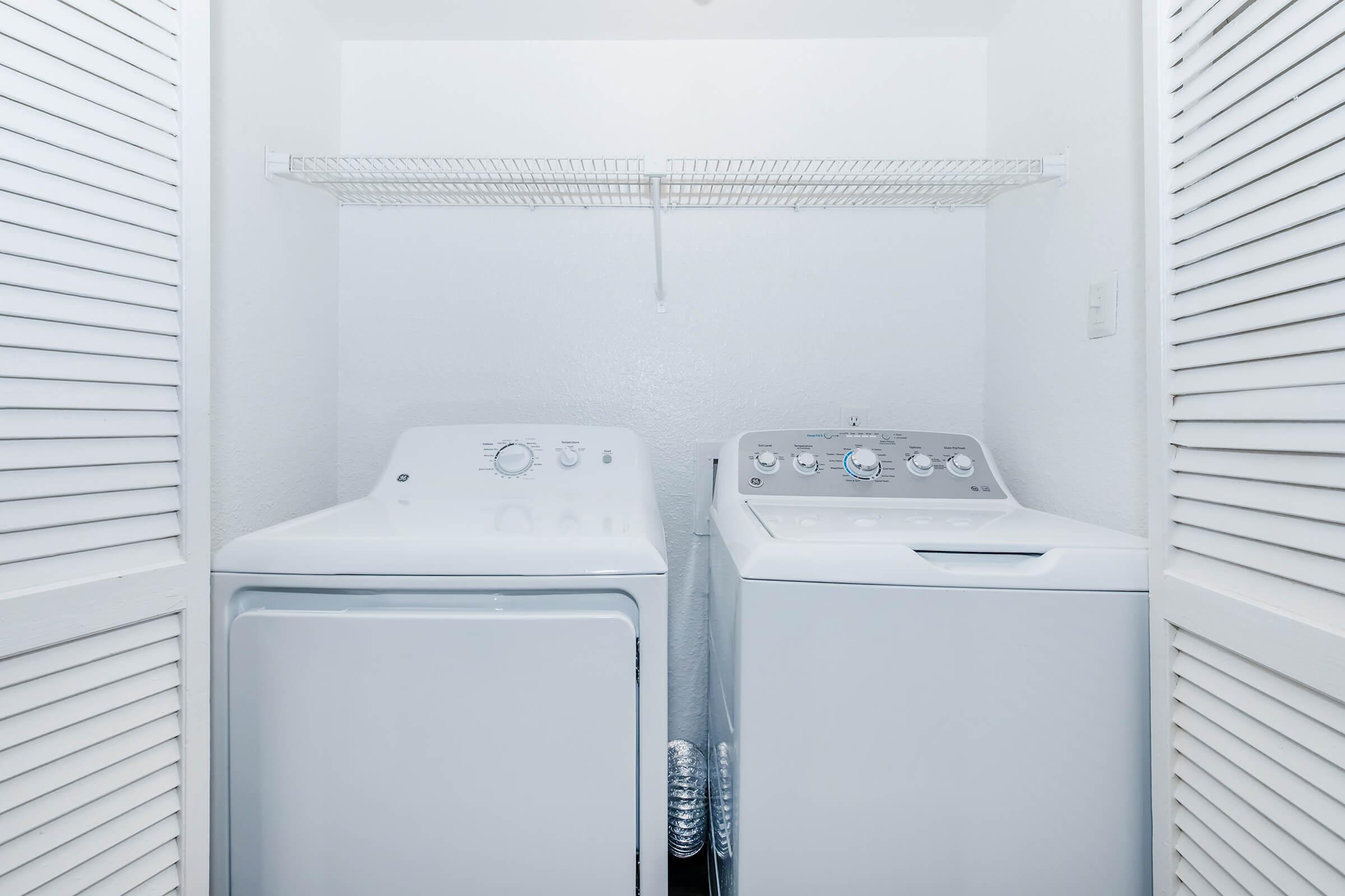 A clean laundry room featuring a white washing machine and a matching dryer side by side, with a metal shelf above for storage. The walls are painted white, and the closet doors are partially open, creating a bright, organized space.