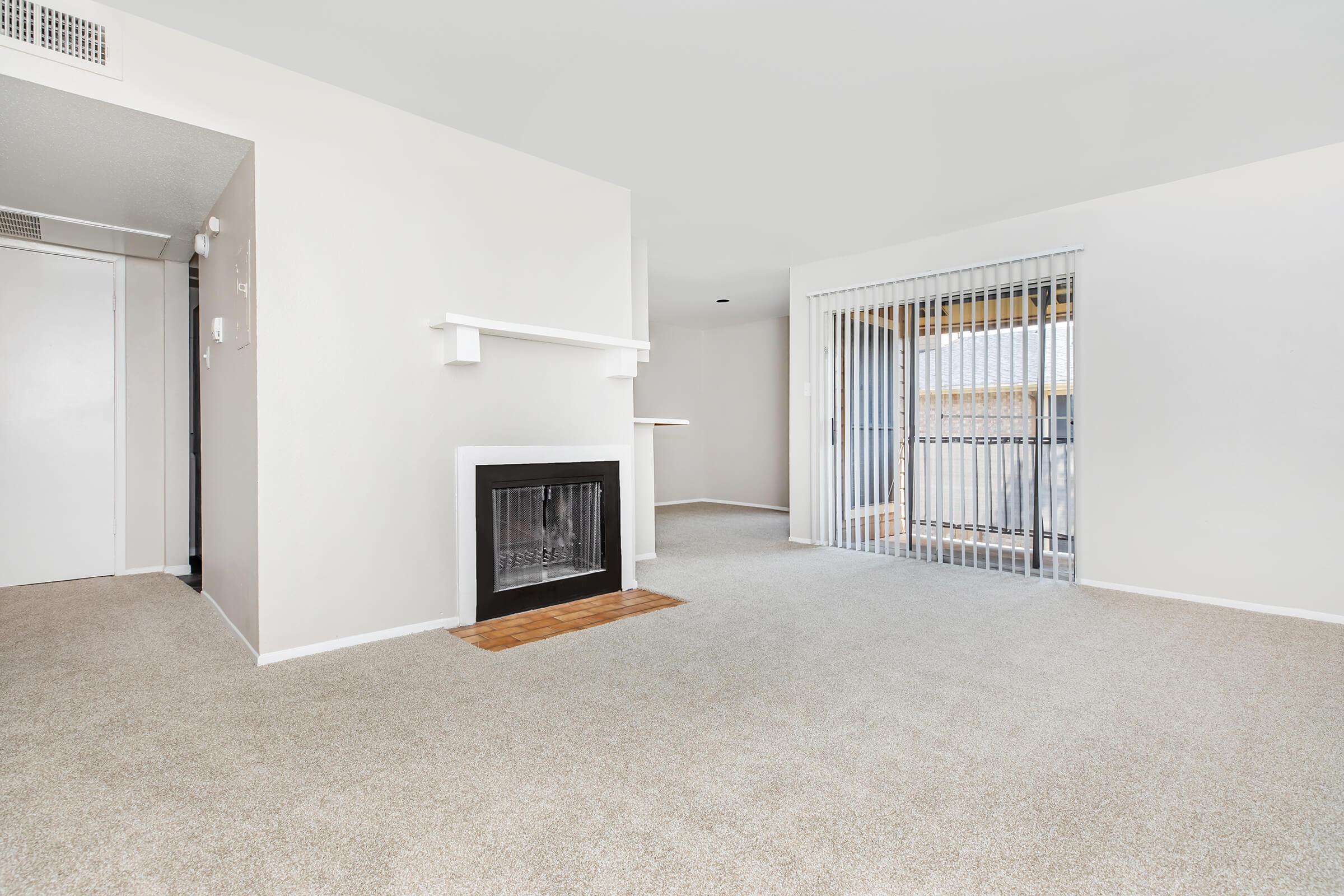 Spacious and bright living room with light-colored walls, plush carpeting, and a decorative fireplace. Large sliding glass door leads to a balcony, allowing natural light to fill the space. The open layout offers a clean and modern aesthetic suitable for various furniture arrangements.