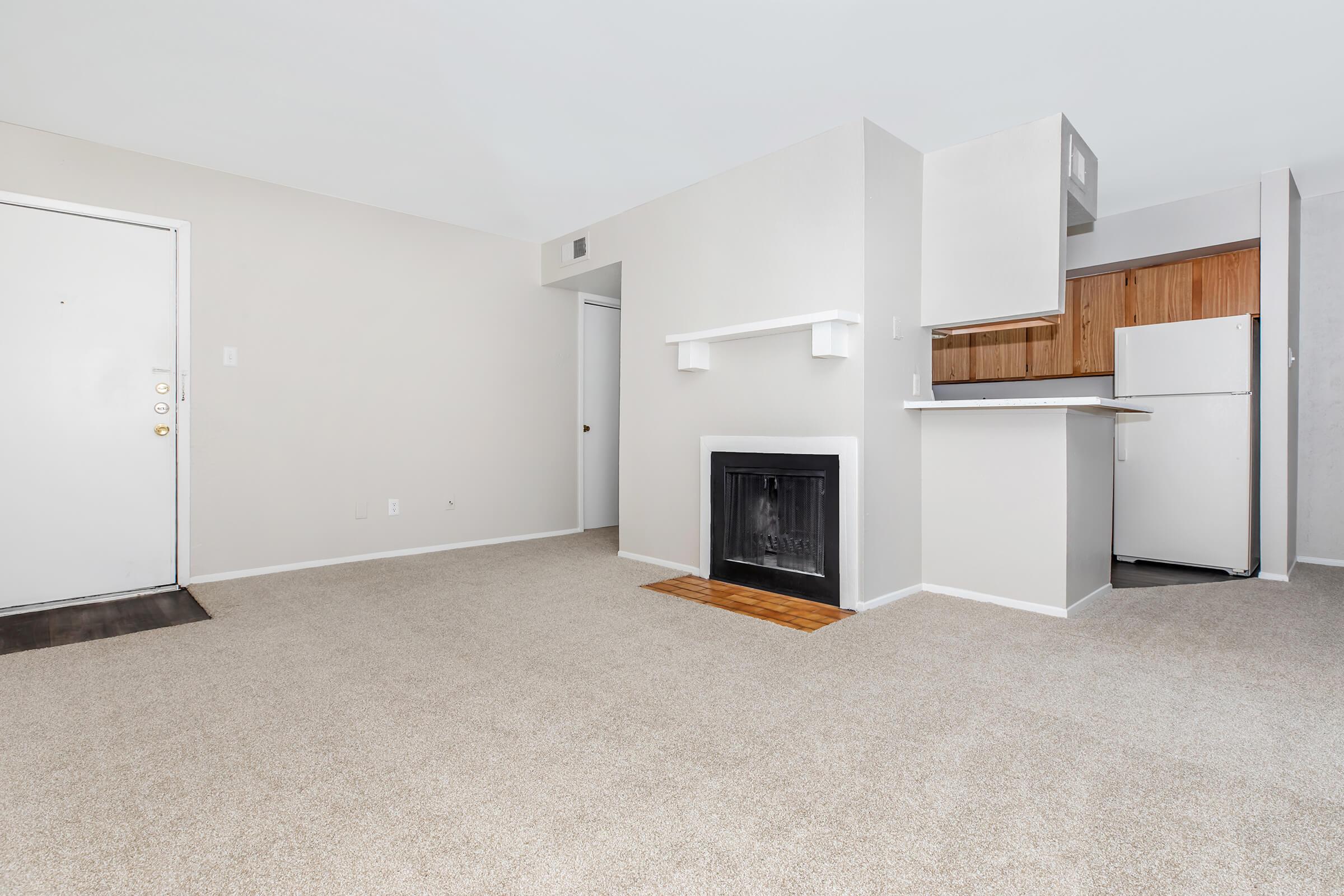 A spacious, empty living room with light beige carpet and neutral-colored walls. There's a white fireplace against one wall and a doorway leading to another room. A small kitchenette with wooden cabinets and a white refrigerator is visible in the background, creating an open layout.