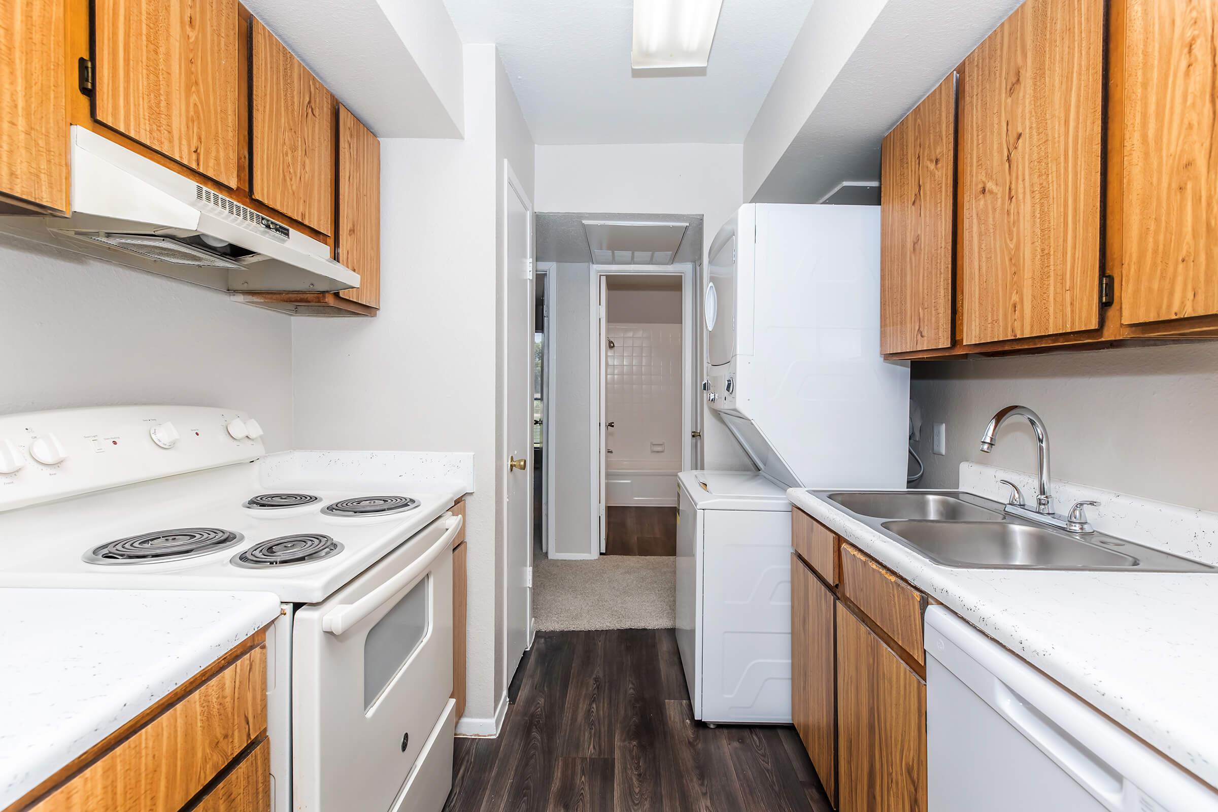 A modern kitchen featuring wooden cabinets, a white stove and refrigerator, a double sink, and a washer/dryer. The walls are painted light, and the flooring is dark. A doorway in the background leads to another room, creating an open and functional space.