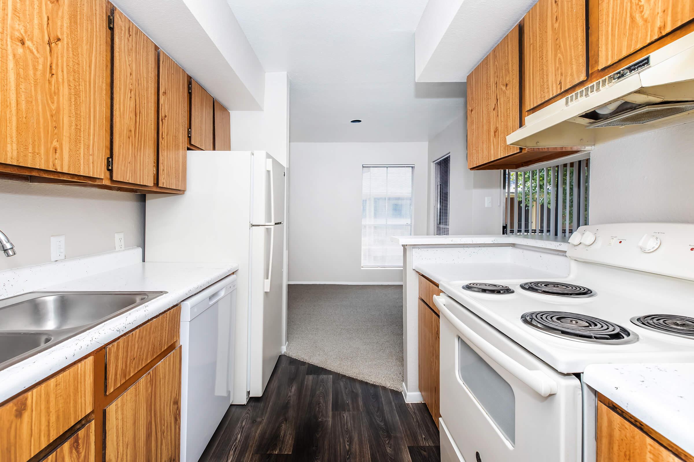 A modern kitchen featuring wooden cabinets, a white stove with burners, a sink, and a refrigerator. The space is bright, with white walls and a view into a carpeted living area with a window. The floor has a dark wood finish, adding contrast to the overall light color scheme.