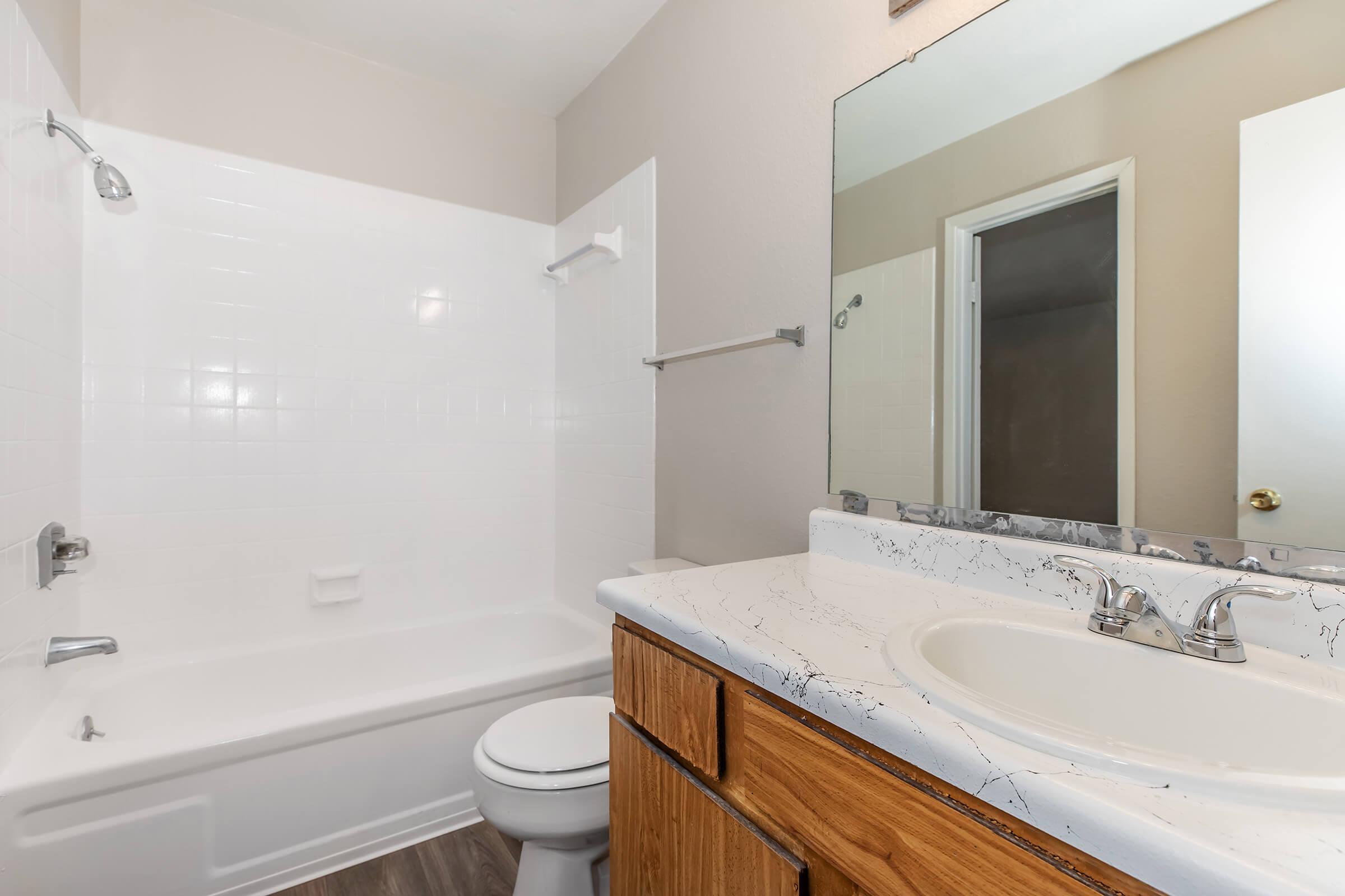 A clean and modern bathroom featuring a white bathtub and shower combination, a wooden vanity with a marble-like countertop, and a large mirror. The walls are painted in a light color, enhancing the bright and spacious feel of the room. A doorway visible in the background leads to another area.