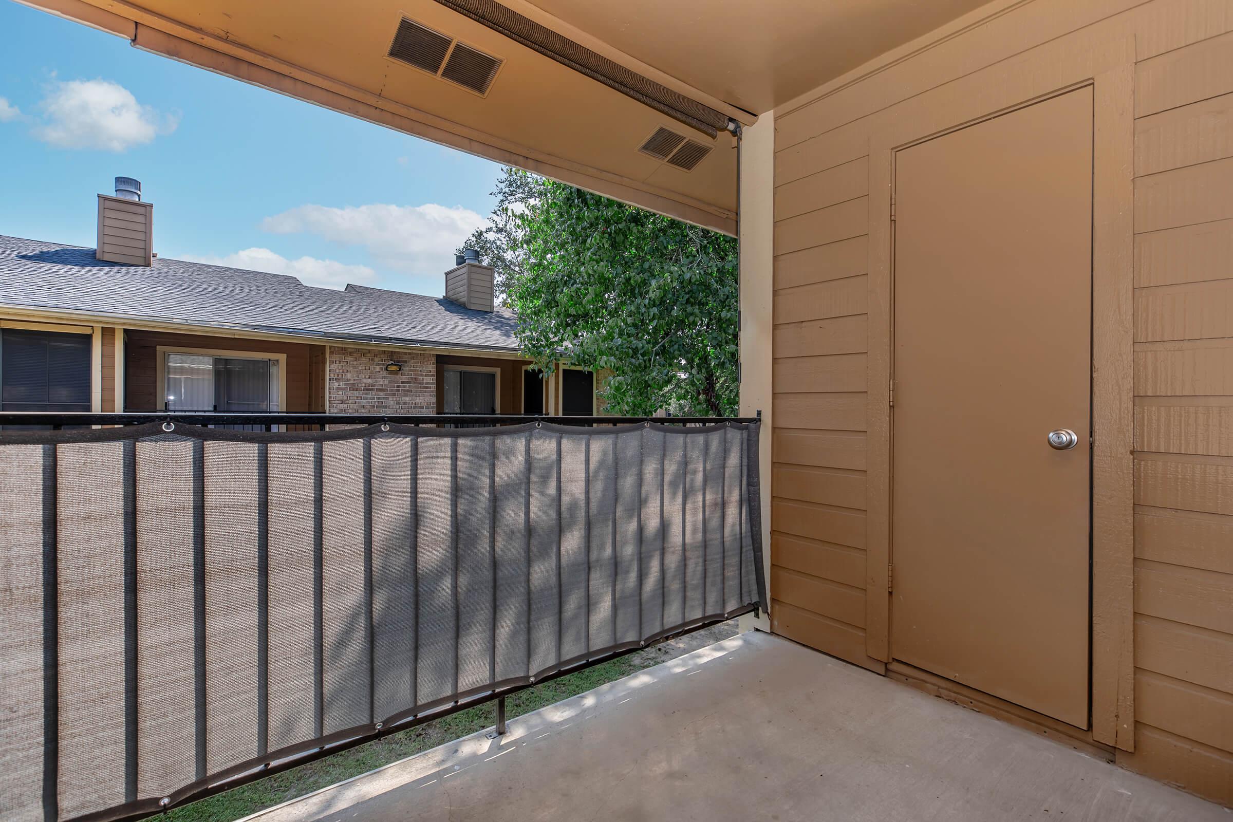 A view of a balcony with a partial railing, featuring a brown door on the left side. In the background, there are other apartments with brown roofs and nearby trees, under a clear blue sky. The concrete floor of the balcony is visible, along with some greenery below.
