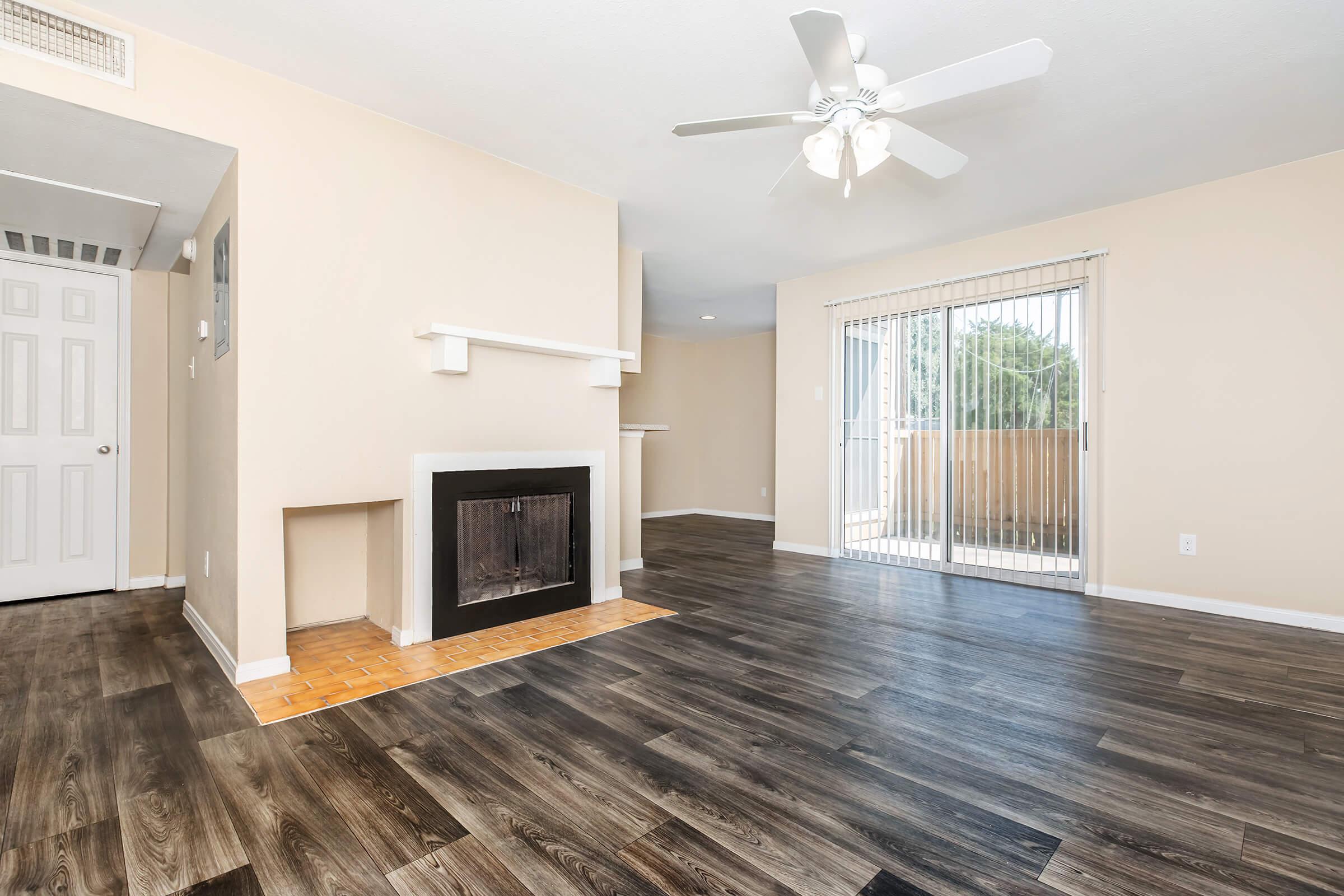 A bright living room featuring light beige walls, dark wood-like laminate flooring, and a ceiling fan. There is a white fireplace with a tiled base, and large sliding glass doors leading to a balcony. The room is well-lit and spacious, creating a welcoming atmosphere.