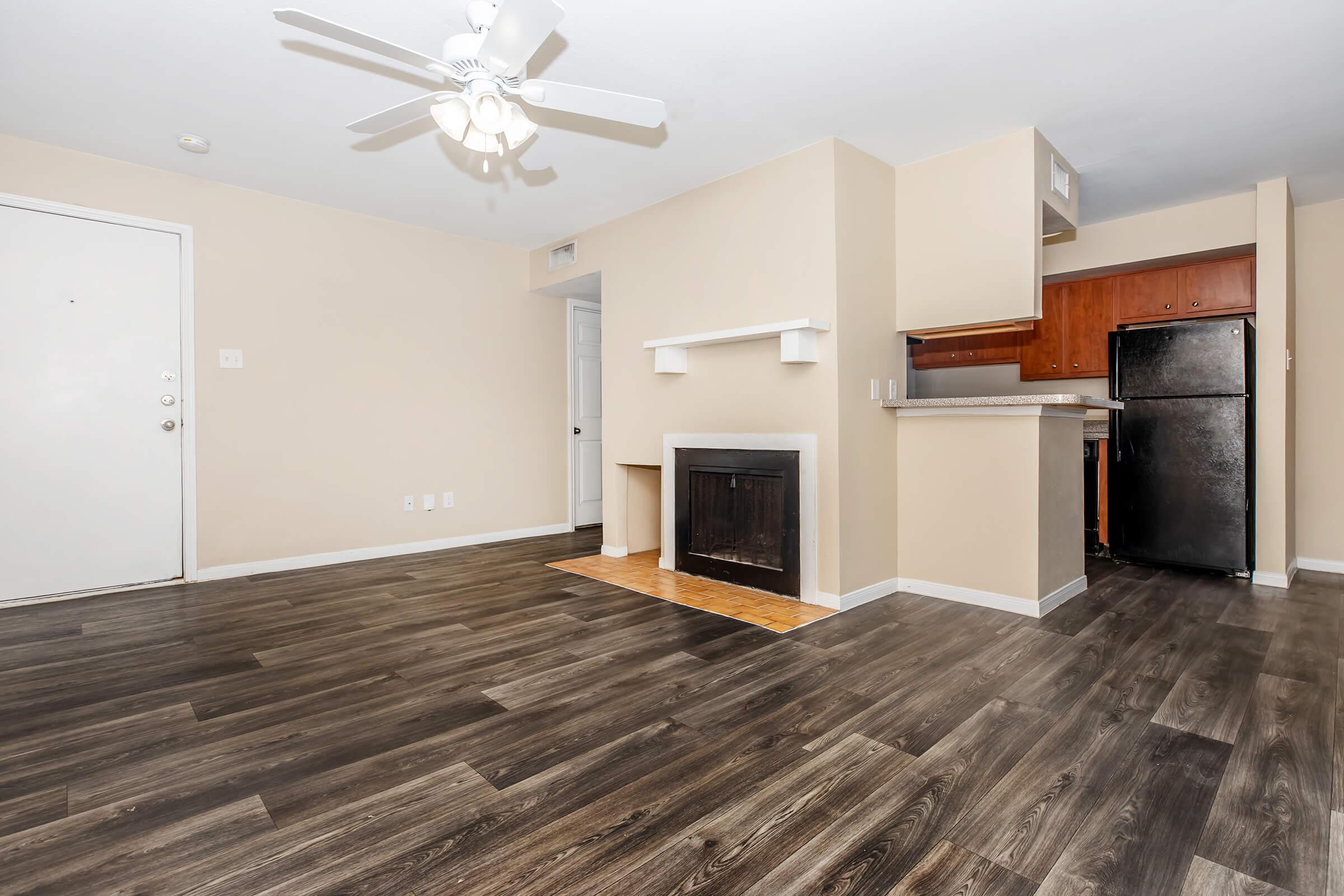 Spacious interior of an apartment featuring a beige wall color, wood-like flooring, ceiling fan, and a decorative fireplace. The kitchen area shows dark wooden cabinets and a black refrigerator in the background. Bright, open layout with natural light streaming in from a window.