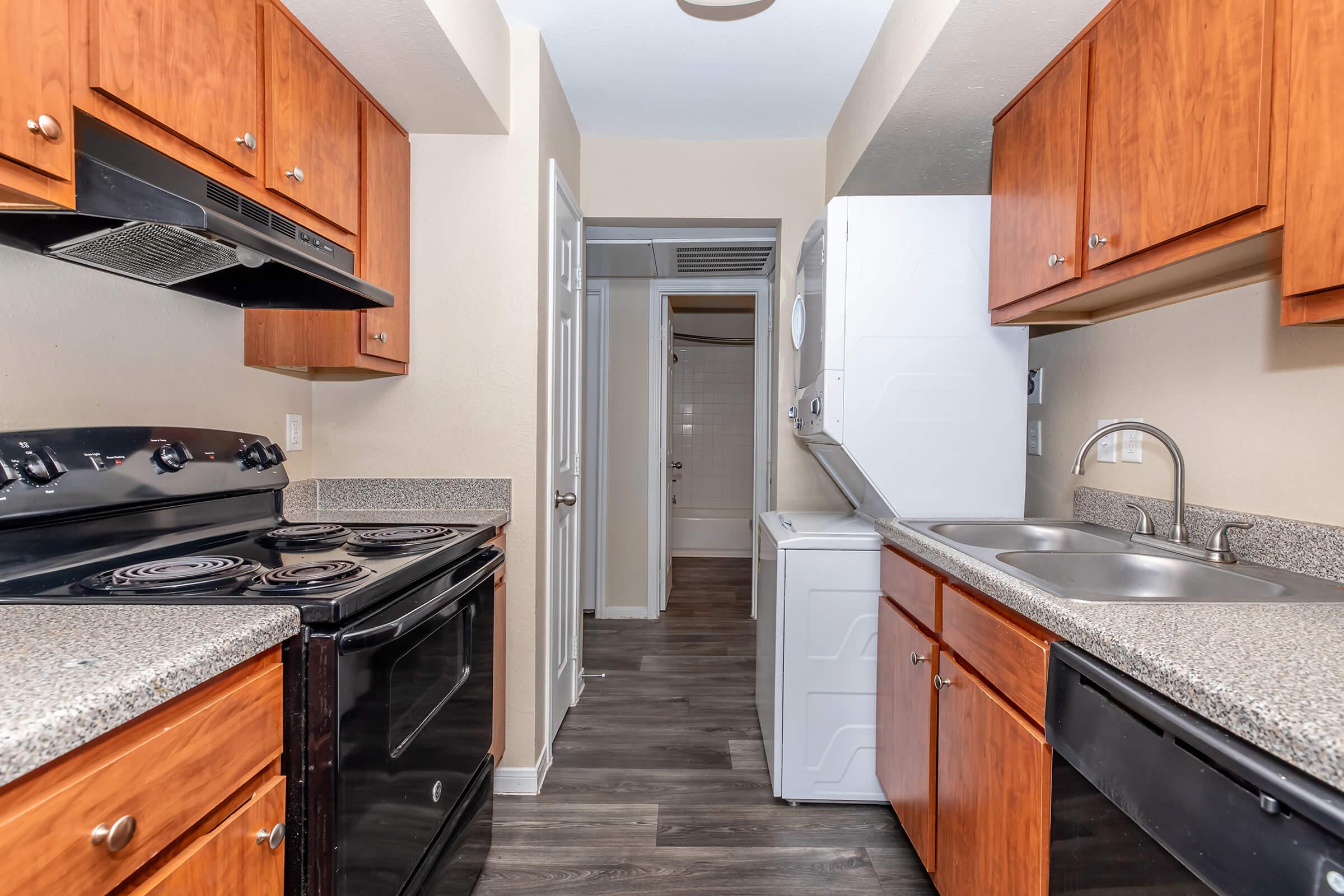 A modern kitchen featuring wooden cabinets, granite countertops, a black stove, and a washing machine. The kitchen is well-lit with neutral wall colors and a clear view into a laundry area. The flooring is dark, adding contrast to the lighter cabinetry.