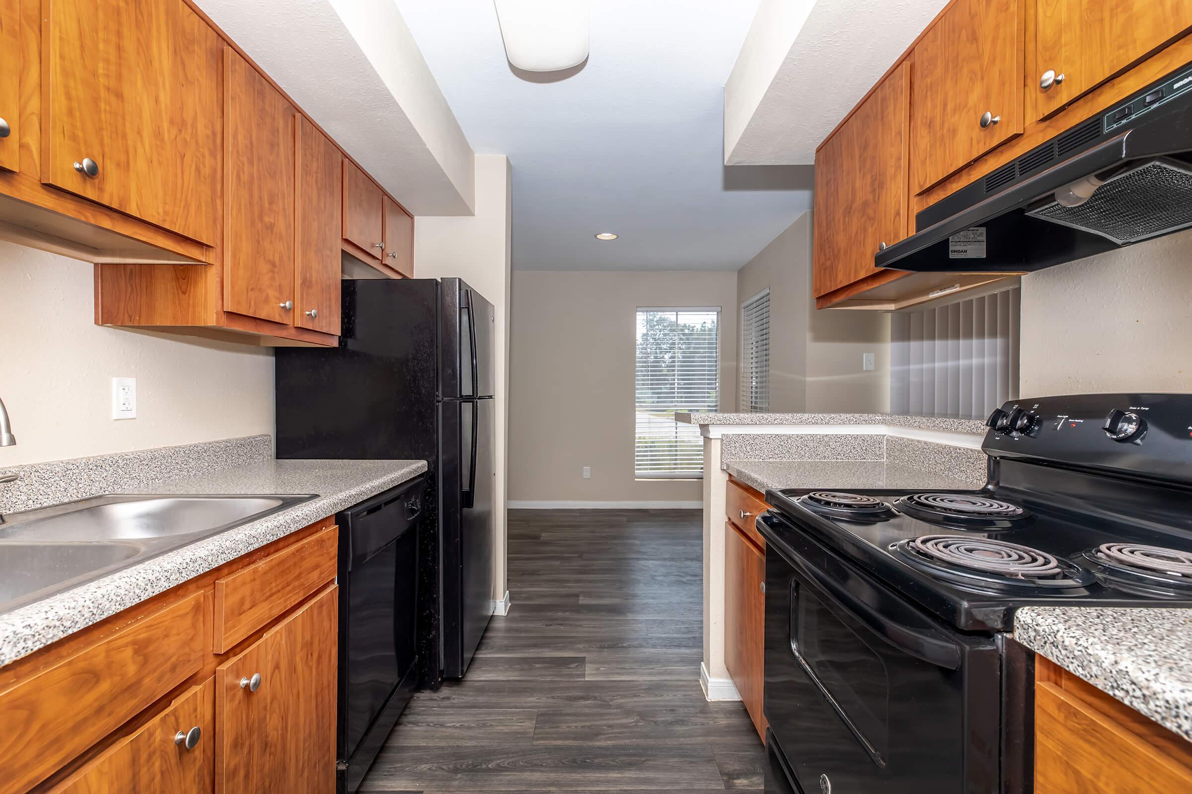 A modern kitchen featuring wooden cabinets, a black refrigerator, and an electric stove. The countertops are gray speckled, and the flooring is dark. A window is visible in the background, allowing natural light into the space.