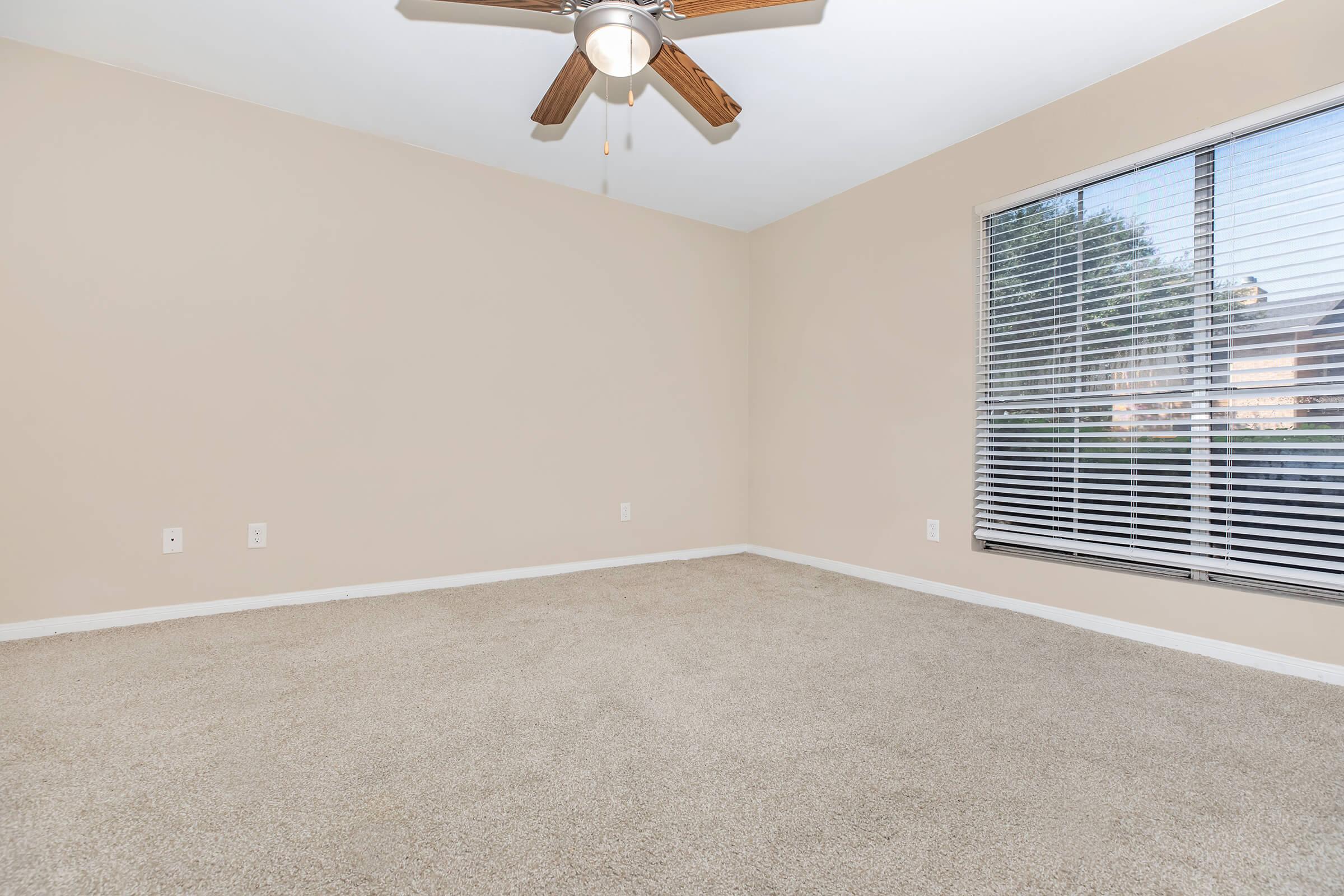 Empty room with beige walls and light brown carpet. A ceiling fan with wooden blades hangs from the ceiling. There is a large window with blinds allowing natural light to enter, alongside a view of the outside space. The room is uncluttered and spacious, ideal for various uses.