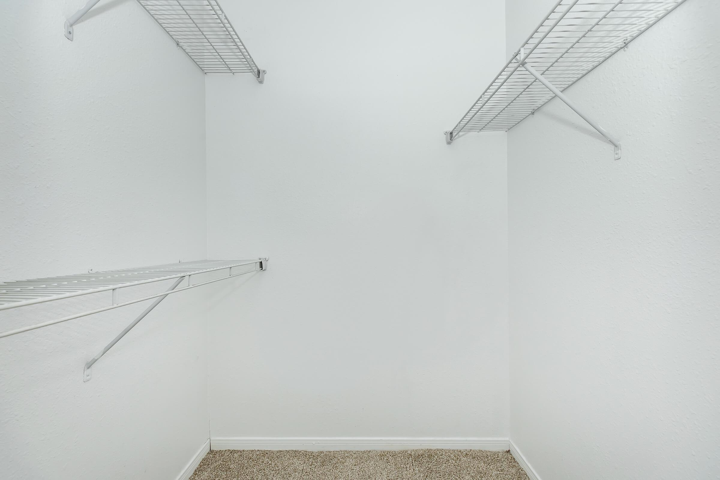 A spacious, empty closet with white walls and a carpeted floor. Two wire shelves are mounted on opposing walls, offering storage options. The overall atmosphere is clean and minimalistic, suitable for organizing clothes or other items.