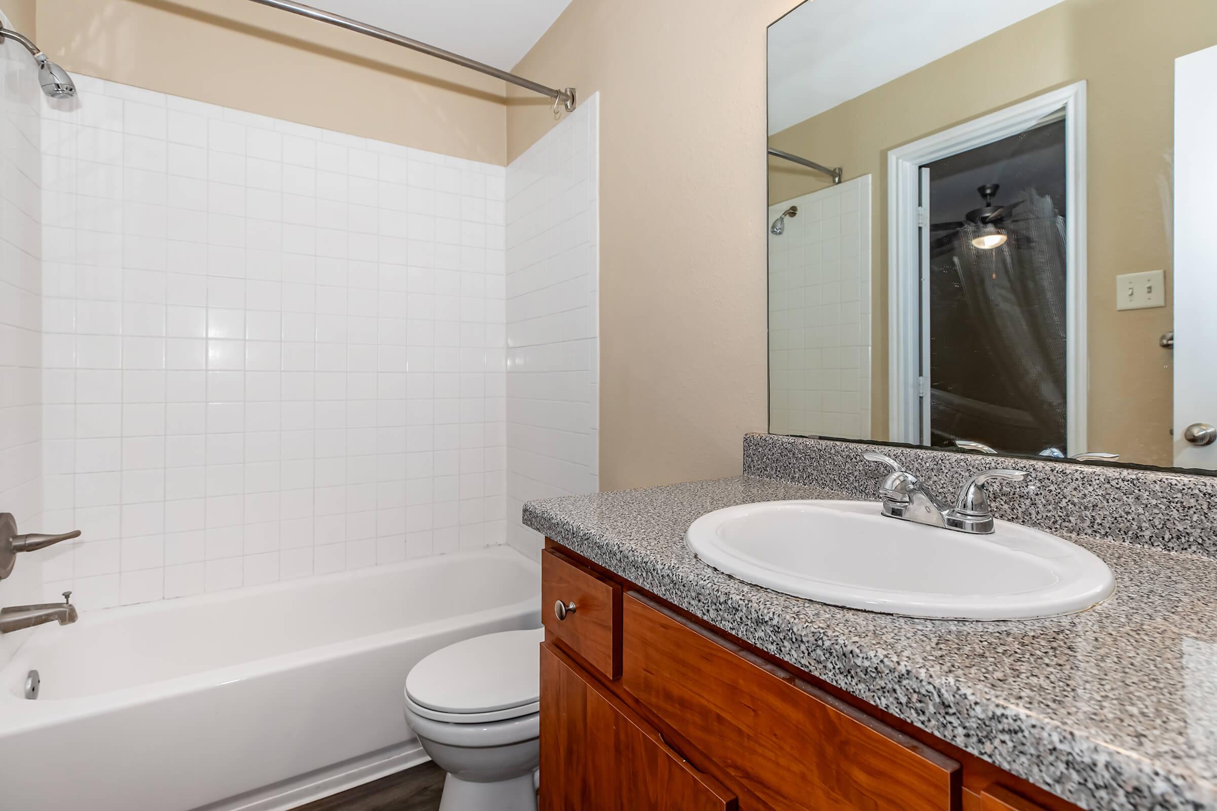 A bright bathroom featuring a white tiled shower and bathtub combo, a wooden vanity with a sink and granite countertop, and a large mirror reflecting the room. The walls are painted a light color, and there is a door leading to another room. The flooring appears to be a dark laminate.