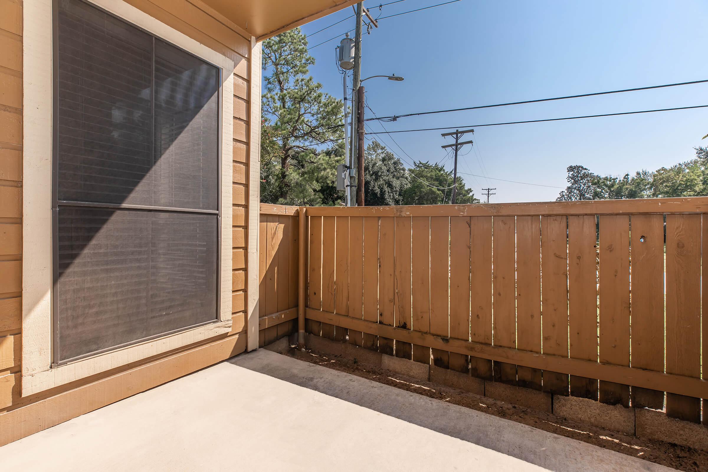 A small outdoor patio with a wooden fence and a concrete floor. There is a window visible on one side, with trees and utility poles in the background under a bright blue sky.