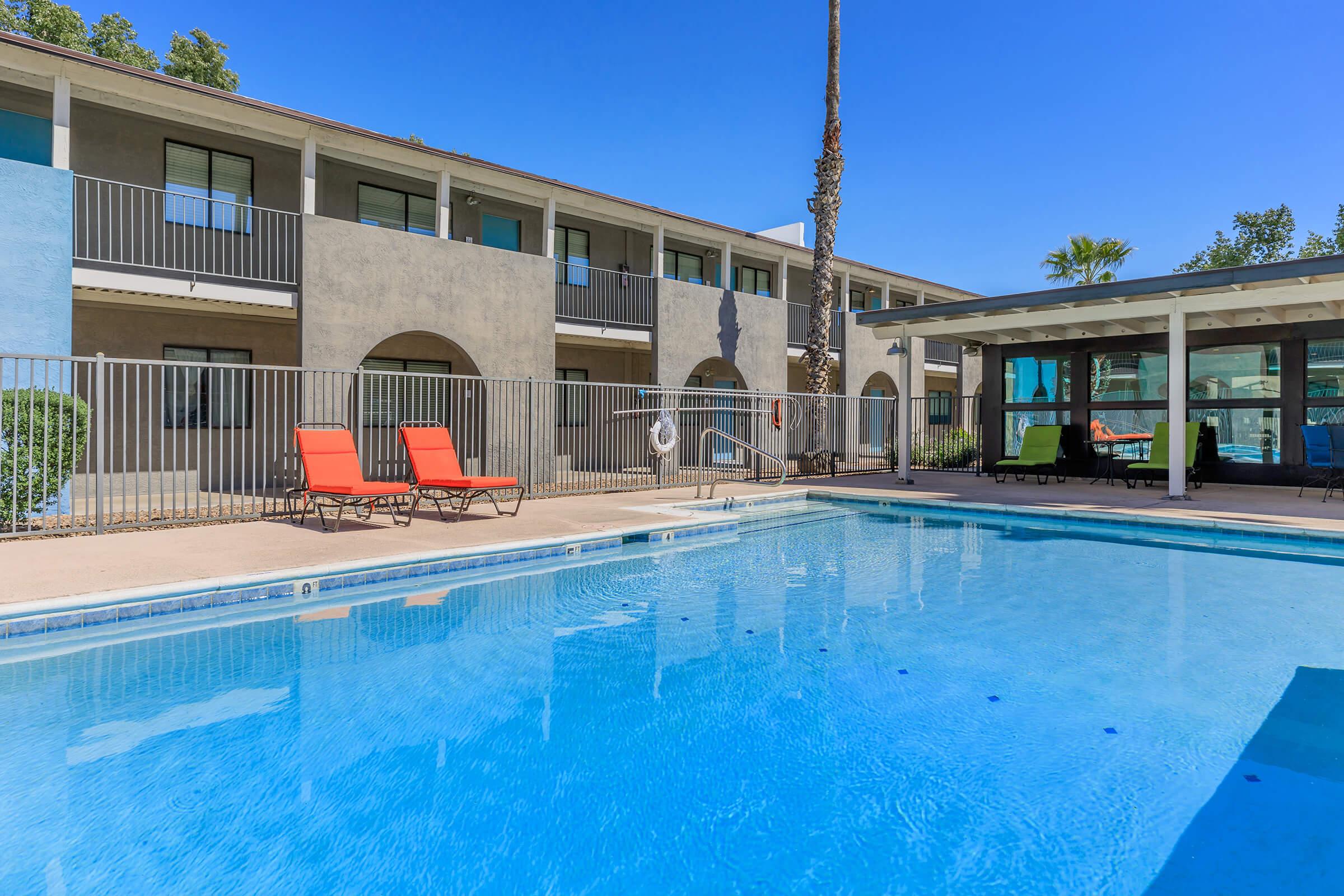 A clear blue swimming pool with lounge chairs in the foreground, surrounded by a beige building with balconies. The area is sunny, with a few palm trees visible. There is a covered seating area nearby, creating a relaxing atmosphere.