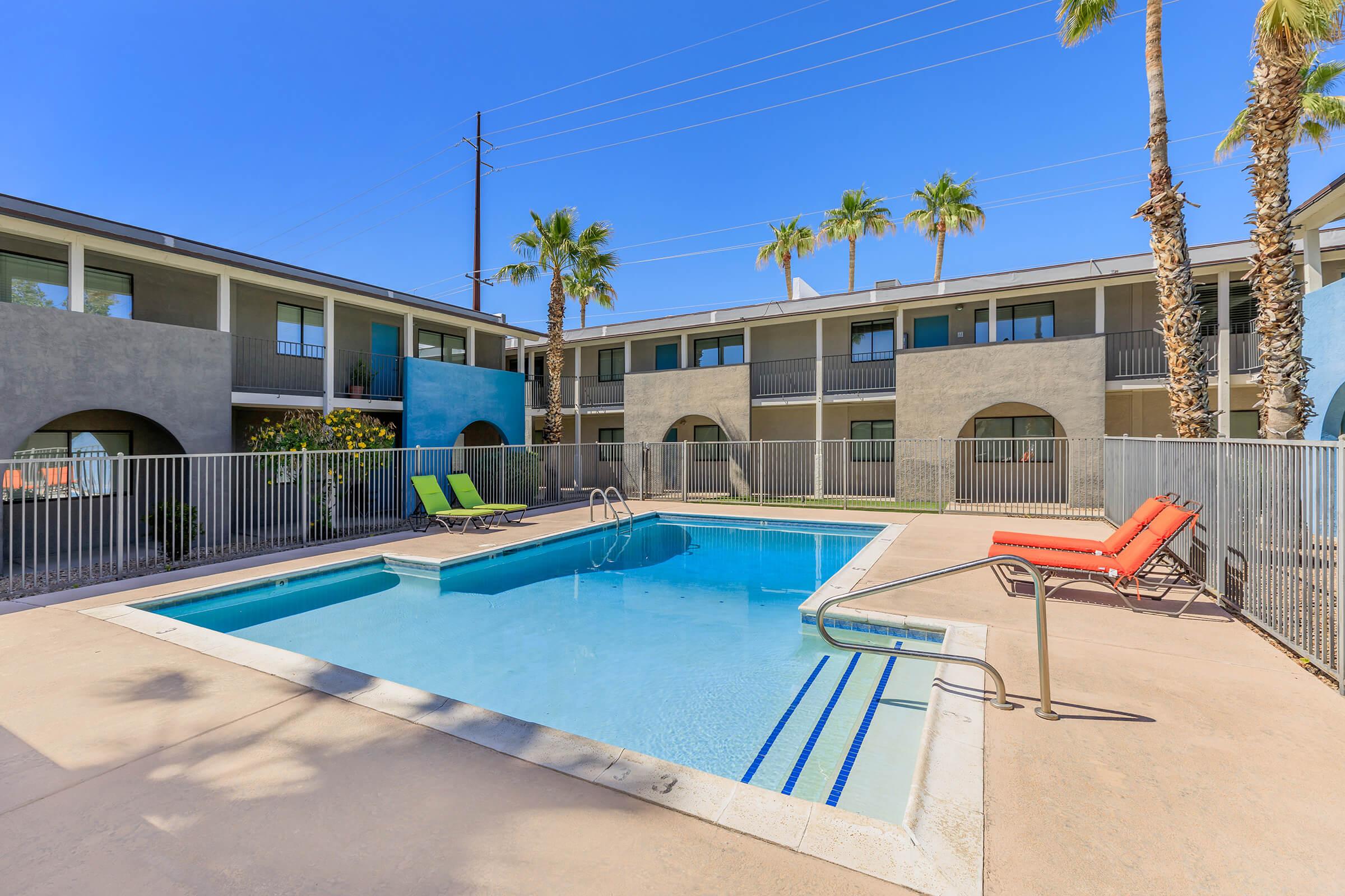 A sunny pool area surrounded by palm trees, with two lounge chairs—one green and one orange—on the pool deck. The pool features clear blue water and is bordered by a low fence. In the background, two-storied apartments with balconies can be seen against a clear blue sky.