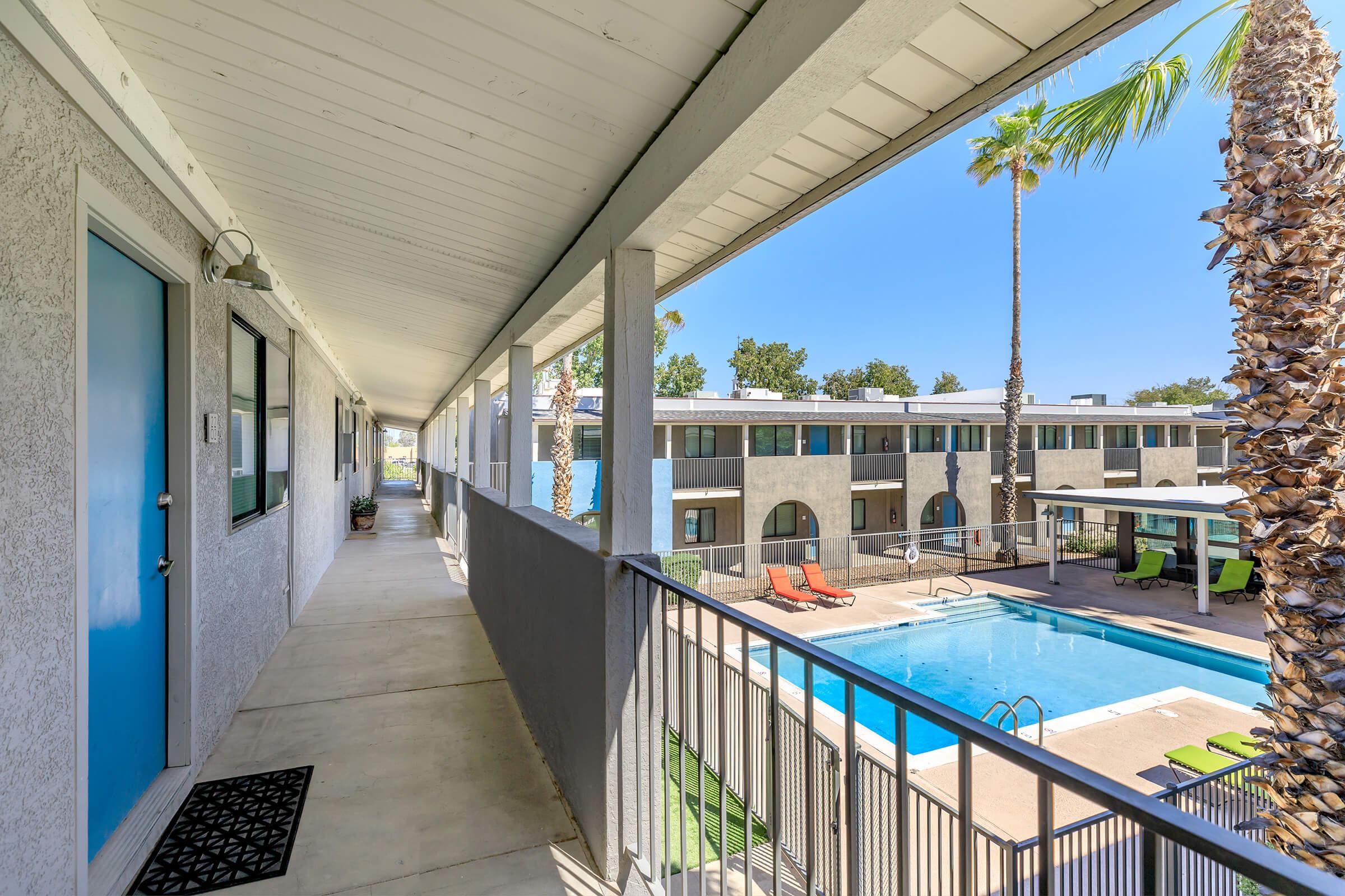 View of a hallway leading to a pool area in an apartment community. The walkway features a blue door on the left, with palm trees and a swimming pool visible in the background. Sunlight creates a bright and inviting atmosphere around the poolside lounge chairs.