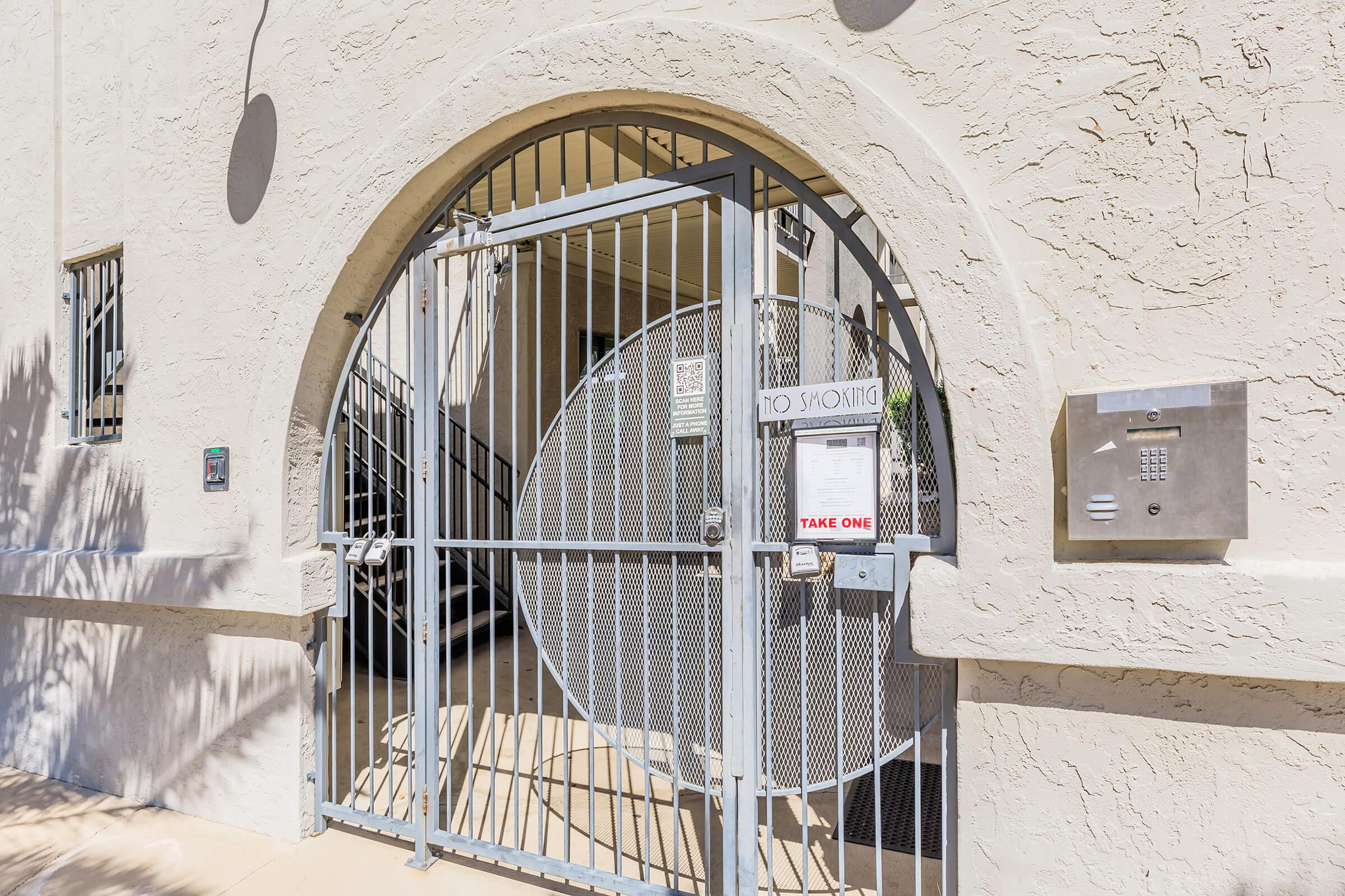 A gated entrance featuring a circular design, with a lockbox on the right side. The gate is painted in a light color and there are signs indicating rules or information. The surrounding wall is textured in a light shade, and sunlight casts shadows in the area.