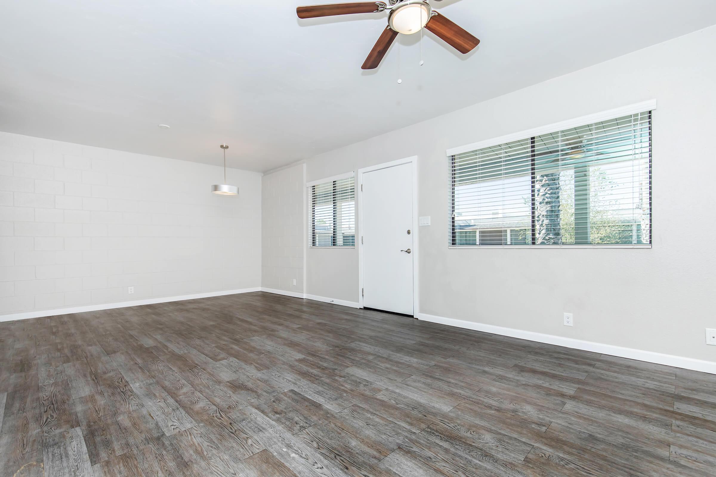 A spacious, empty room with gray walls and a wooden floor. There is a ceiling fan with wooden blades, a pendant light fixture, and large windows with blinds letting in natural light. A white door leads to another area, creating an open and bright environment.