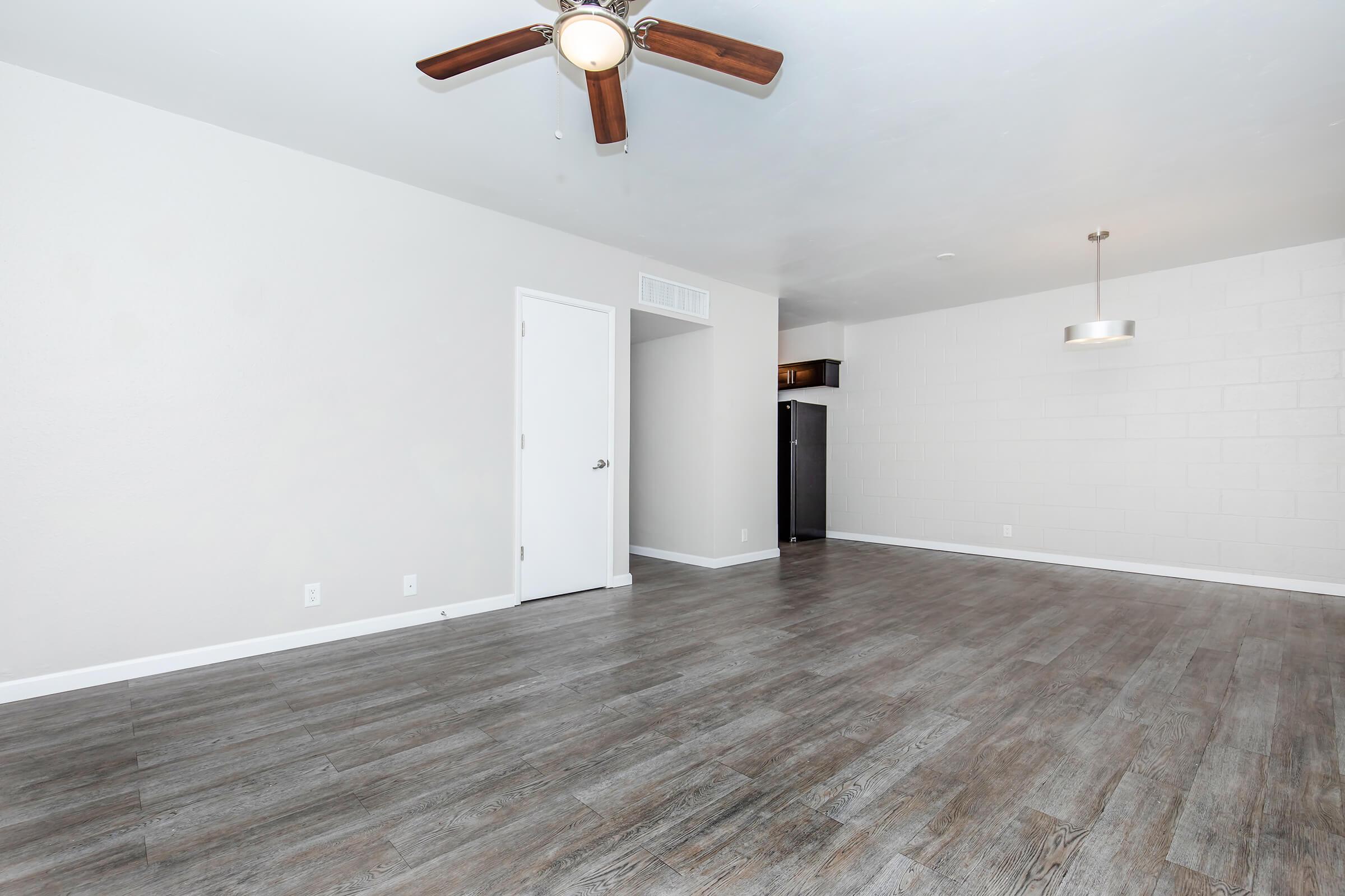 Spacious, well-lit living area with light-colored walls and wood laminate flooring. A ceiling fan is installed in the center, and a doorway leads to another room. In the background, there is a modern pendant light and a dark kitchen area with a refrigerator visible.