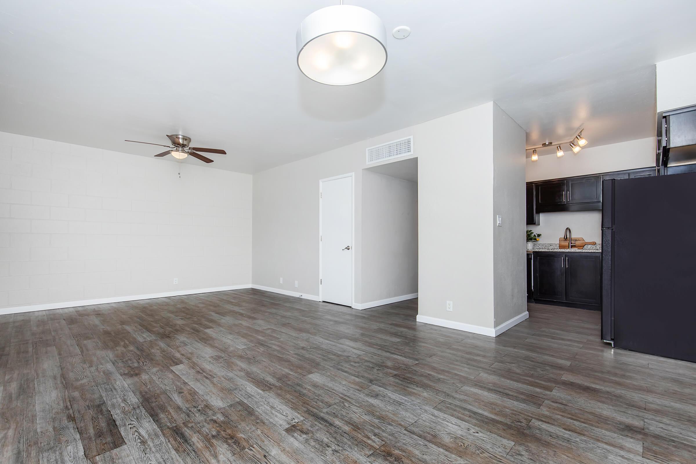 A spacious, modern living area with light-colored walls and a ceiling fan. The room features hardwood-style flooring, a large window, and a doorway leading to a kitchen area with dark cabinets. Soft lighting fixtures enhance the bright atmosphere. The layout is open and inviting, ideal for various furniture arrangements.