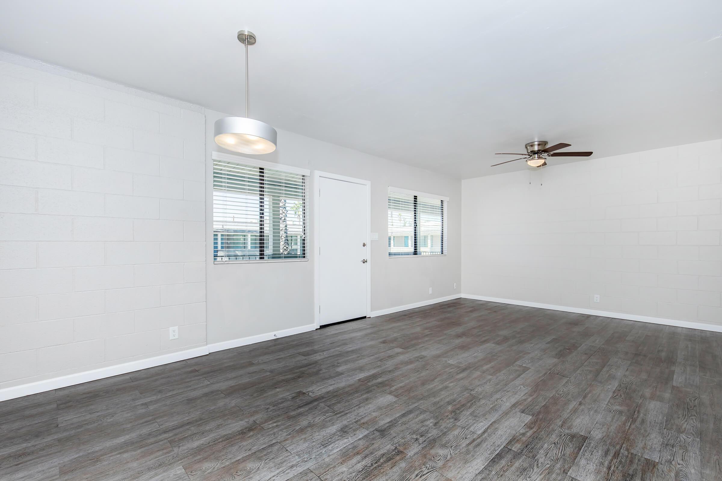 A spacious, empty living room featuring light gray walls and a wood-look floor. There is a ceiling fan with light, a modern light fixture, and large windows allowing natural light. A white door leads outside, creating a bright and airy atmosphere.