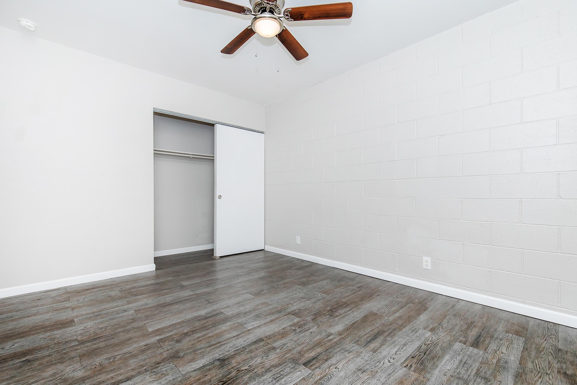 A spacious, empty room with light-colored walls and a wood-like floor. There is a ceiling fan with dark wooden blades, and a closet with a sliding white door is visible. The overall ambiance is bright and minimalist, suitable for different decorating styles.