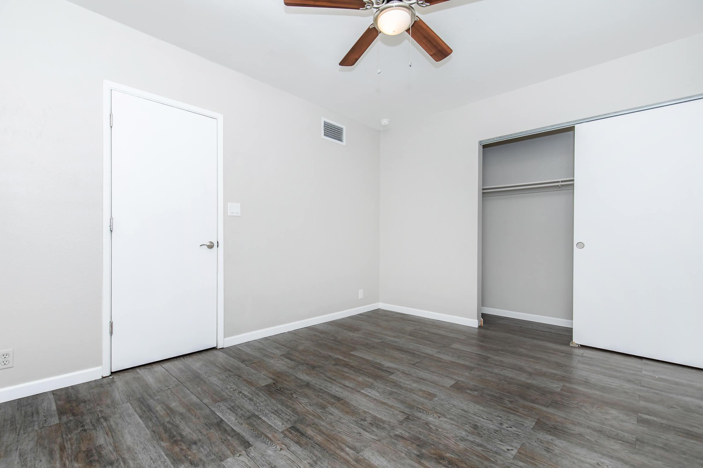 A clean and empty bedroom featuring light-colored walls, a ceiling fan, and a door leading to a closet. The floor is made of wood laminate, and there's a second closet door visible. The overall aesthetic is simple and modern, creating a spacious feel.