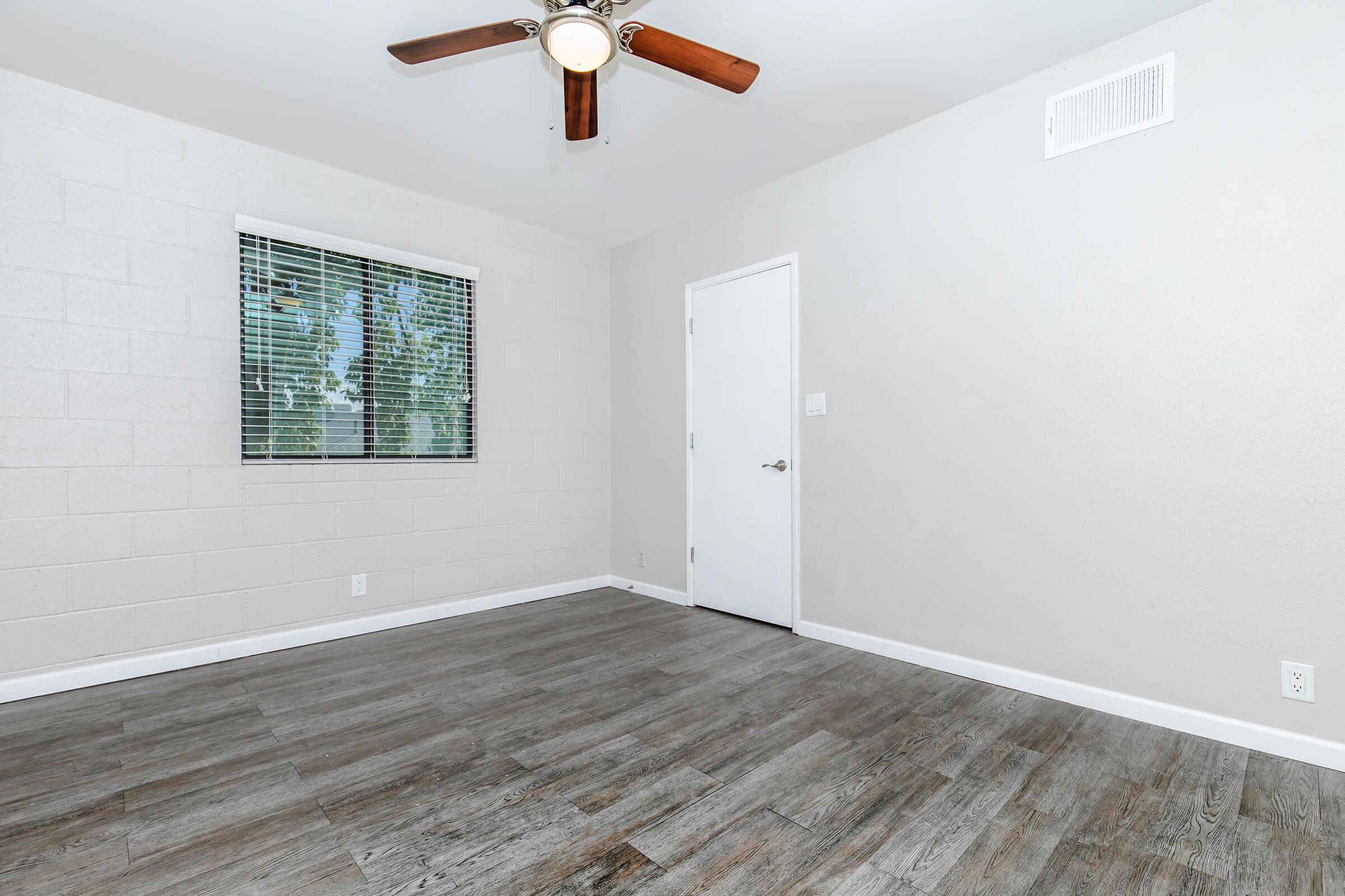 A sparsely furnished room with a ceiling fan and hardwood-like flooring. There is a window with blinds on the left side and a white door on the right, leading to another space. The walls are painted a light gray, creating a clean and modern aesthetic.