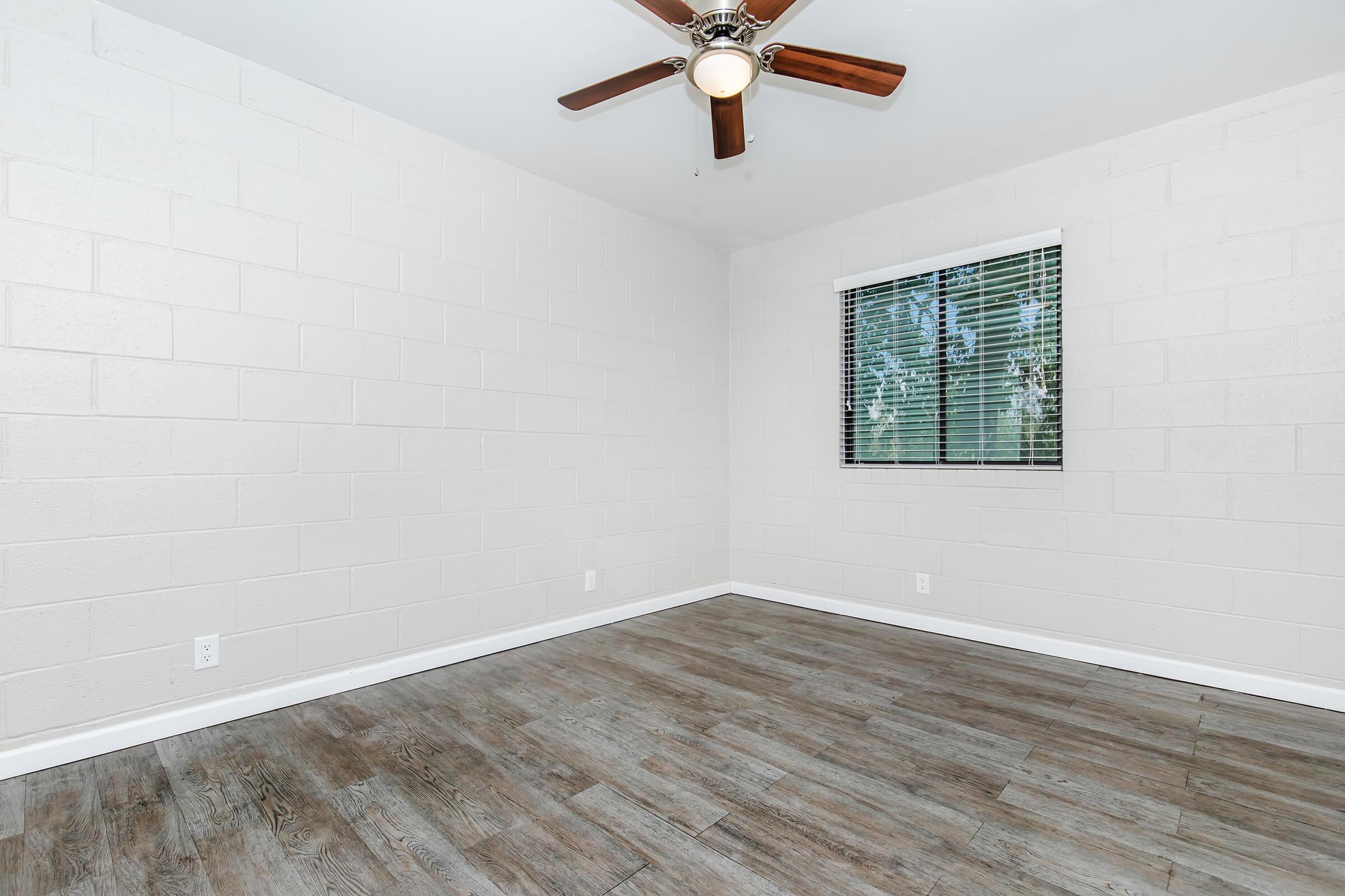 A vacant room with light-colored walls and dark wooden flooring. It features a ceiling fan with wooden blades, and a window with blinds letting in natural light. The space is empty, creating a minimalist and open atmosphere.