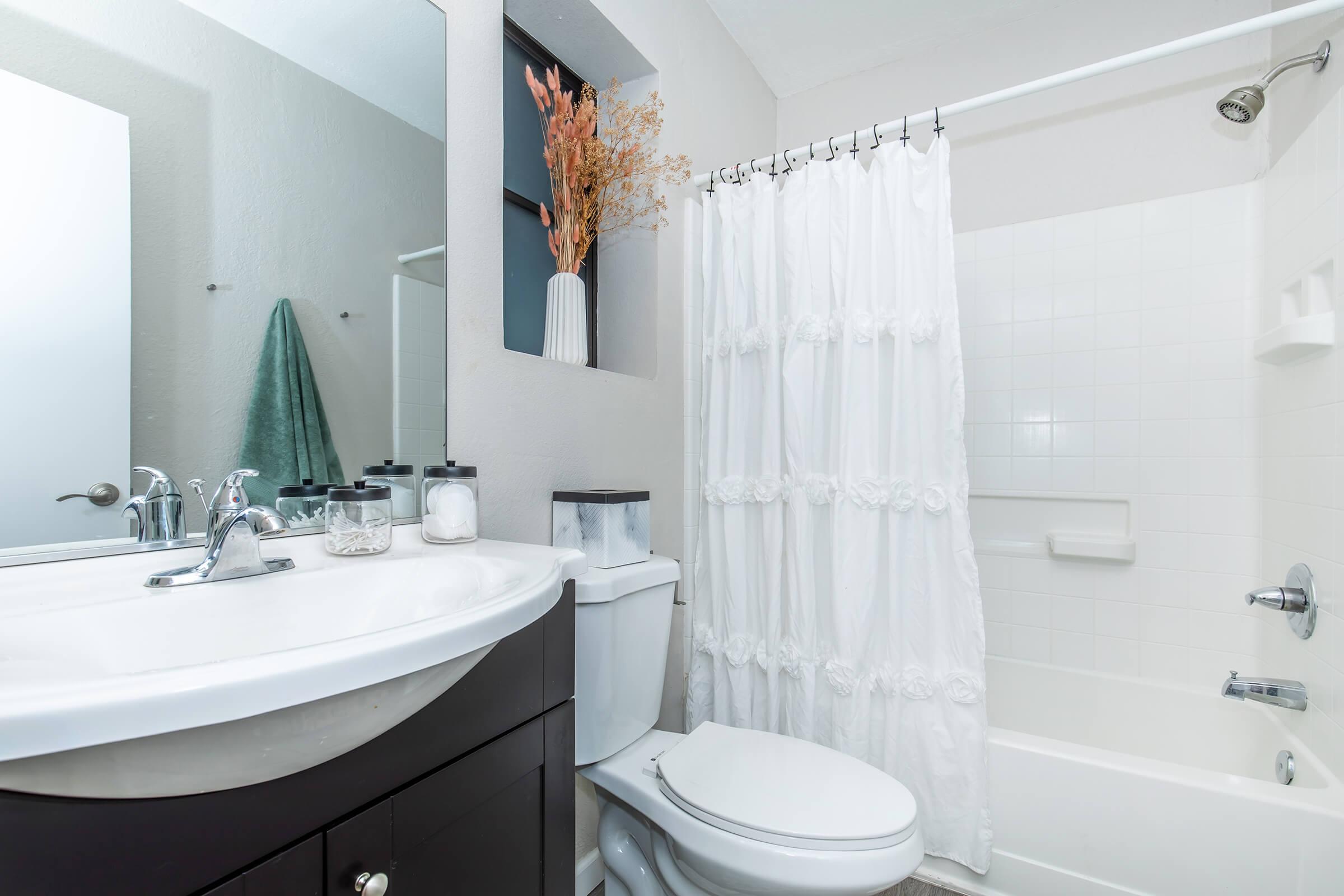 A clean and modern bathroom featuring a white bathtub with a shower curtain, a countertop with a sink and faucets, a wall-mounted mirror, a green towel hanging, and decorative elements like a vase with dried flowers on the shelf. The overall color scheme is neutral with a light and airy feel.