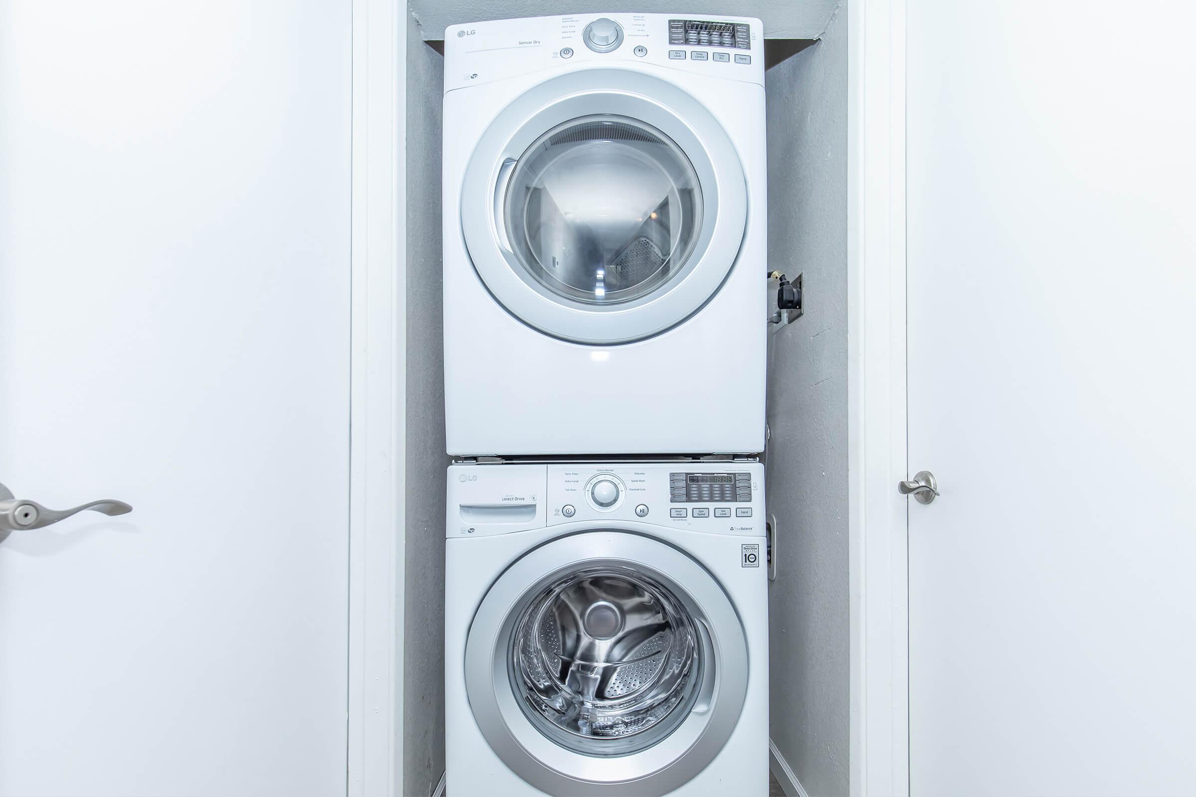 A stacked white washer and dryer located in a narrow laundry space. The washer is on the bottom, featuring a front-loading design with a transparent door. The dryer, positioned above, also has a front-loading design and control panel visible on the front. The walls are painted white, and there is a closed door nearby.