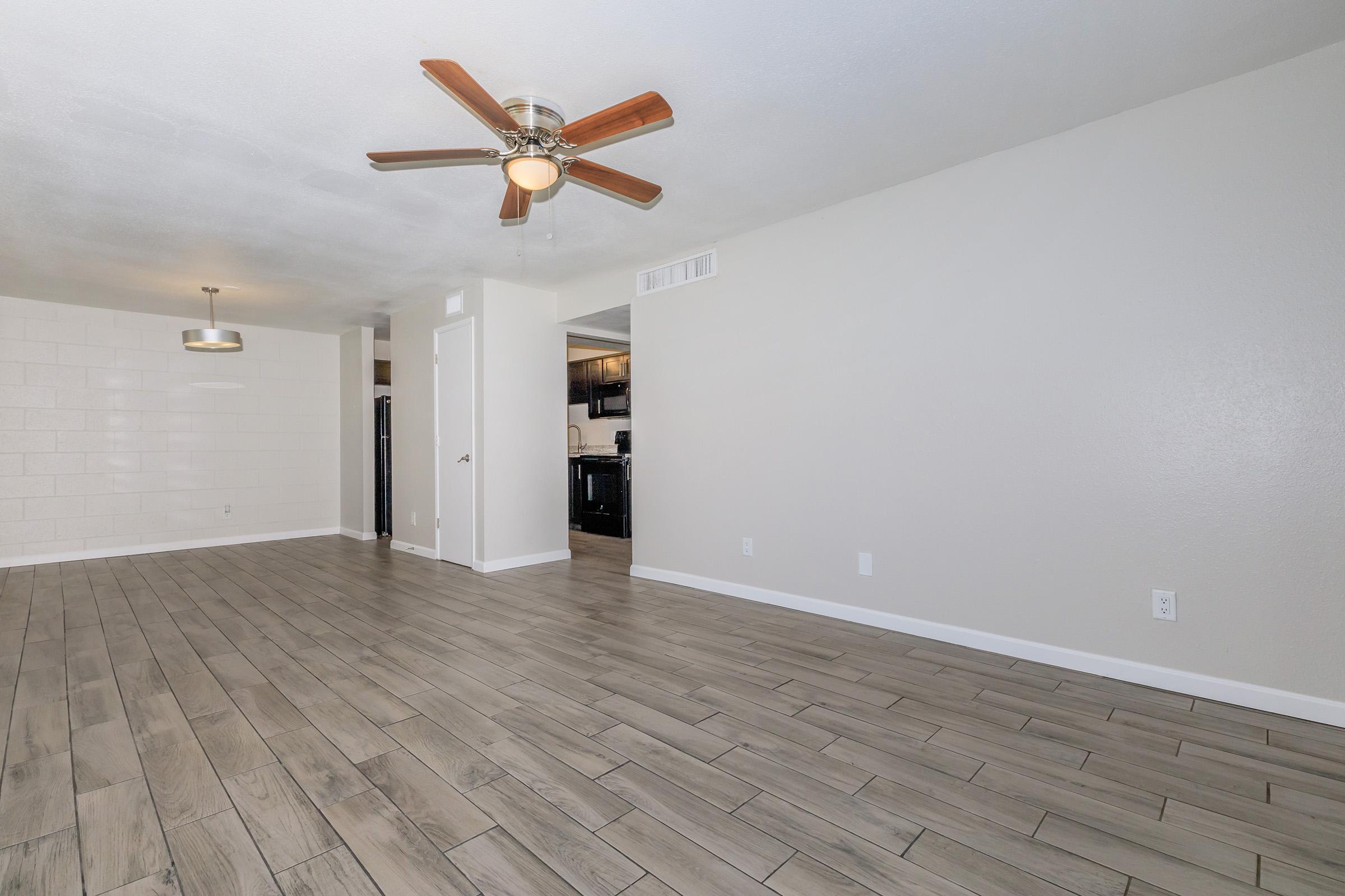 A spacious, empty living room featuring light-colored walls and a modern ceiling fan. The floor is covered in large, rectangular tiles. In the background, a doorway leads to a kitchen area with dark cabinets, and a pendant light hangs above the dining space. The overall atmosphere is bright and inviting.