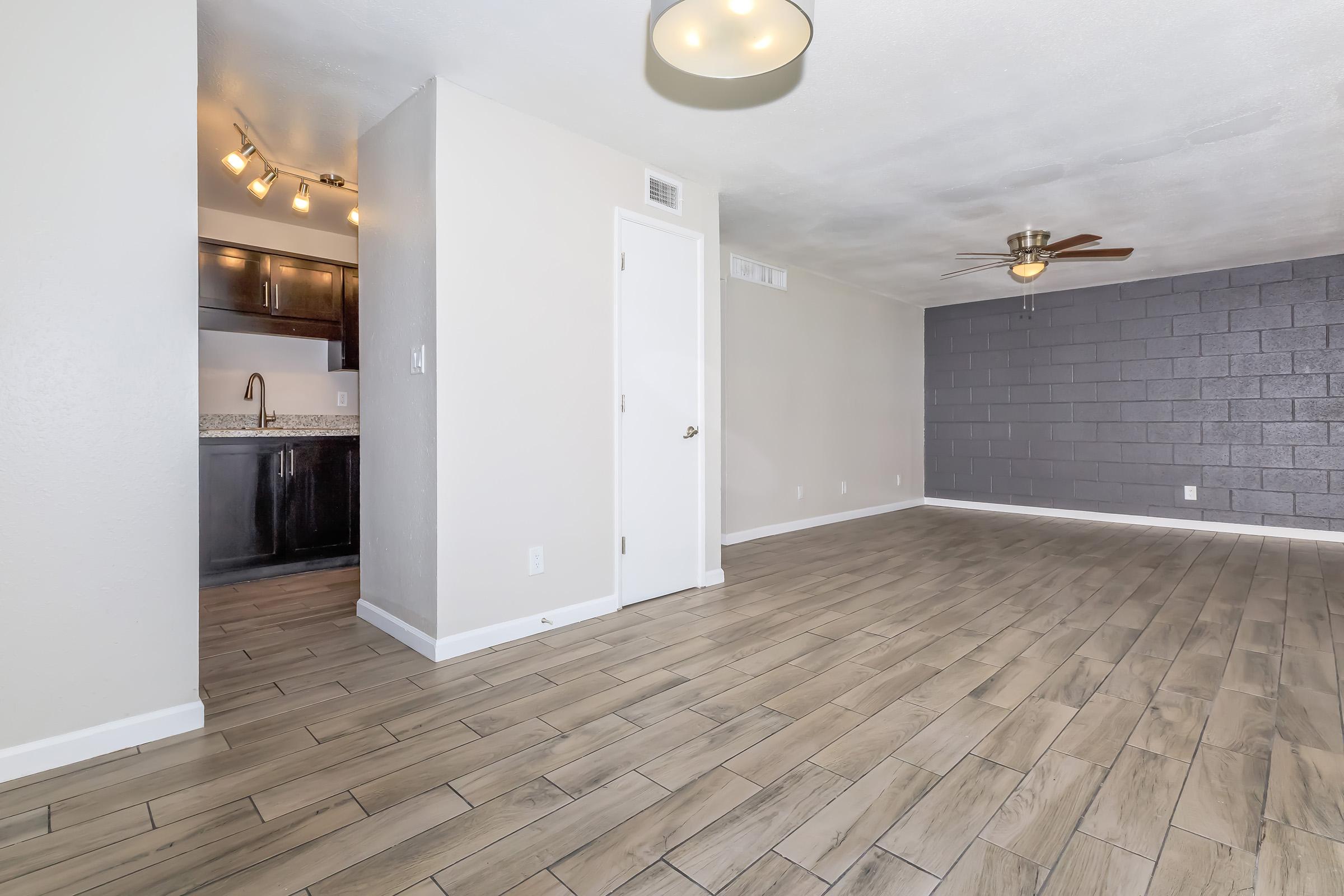 Interior view of a modern, spacious living area featuring light-colored walls, wood-like tile flooring, and a ceiling fan. To the left, an open kitchen area with dark cabinetry and a countertop is partially visible. A gray accent wall adds contrast to the room, which is well-lit by natural and ceiling light.