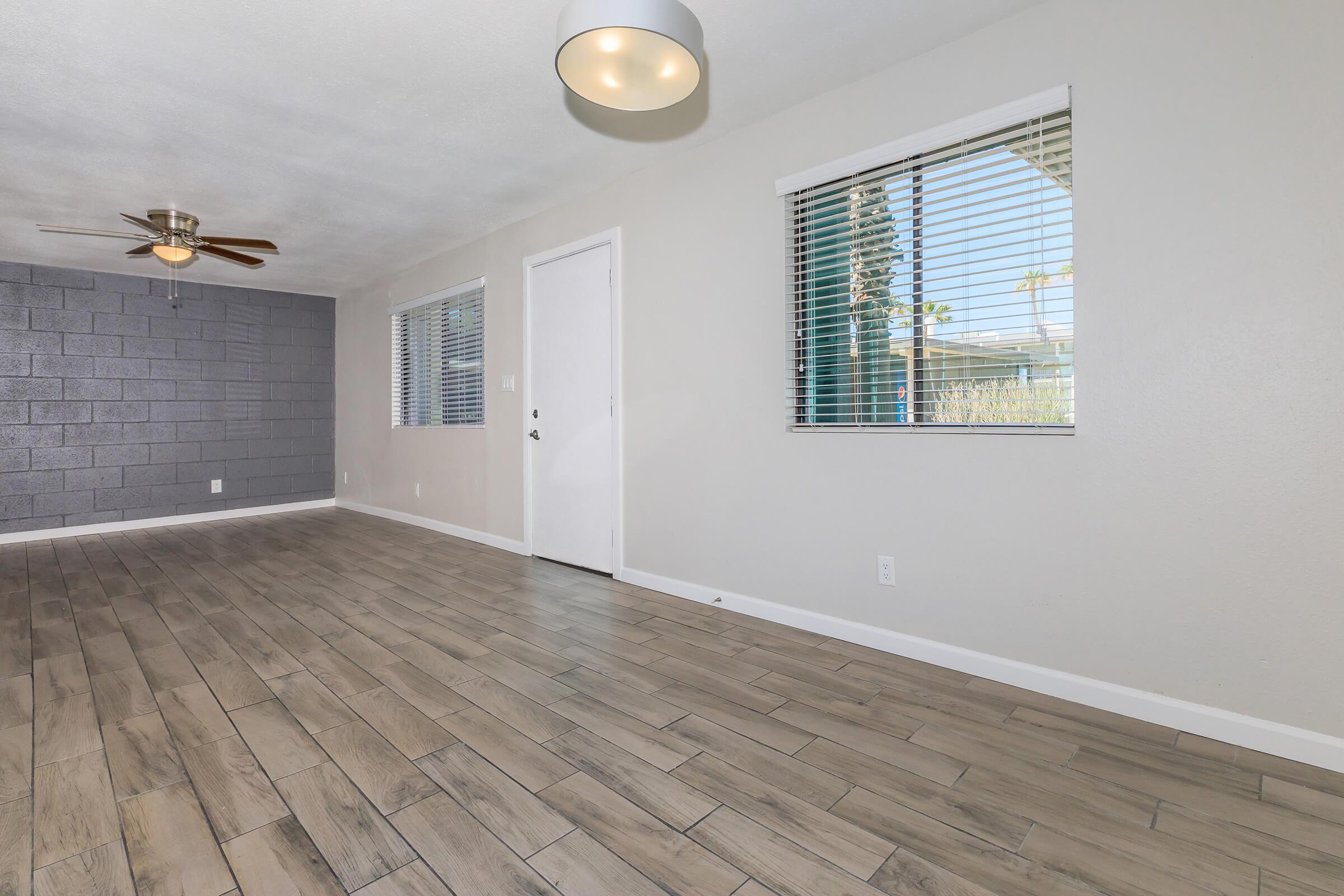 A spacious room featuring light-colored walls, large windows with blinds, and modern tile flooring. There is a ceiling fan on the left and a light fixture hanging from the ceiling. A door leads outside, and one wall is painted in a darker gray hue. The overall ambiance is bright and inviting.