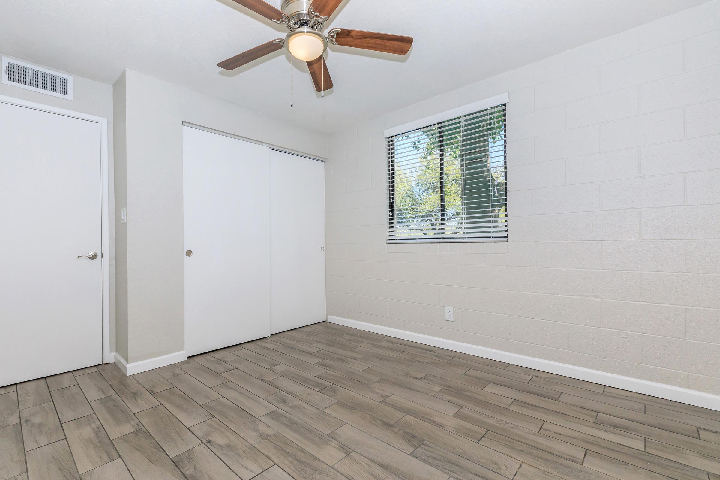 A well-lit room with a ceiling fan, featuring a neutral color palette. There are two white sliding closet doors on one side and a window with blinds allowing natural light. The floor is covered with wood-like tiles, creating a modern, clean atmosphere.