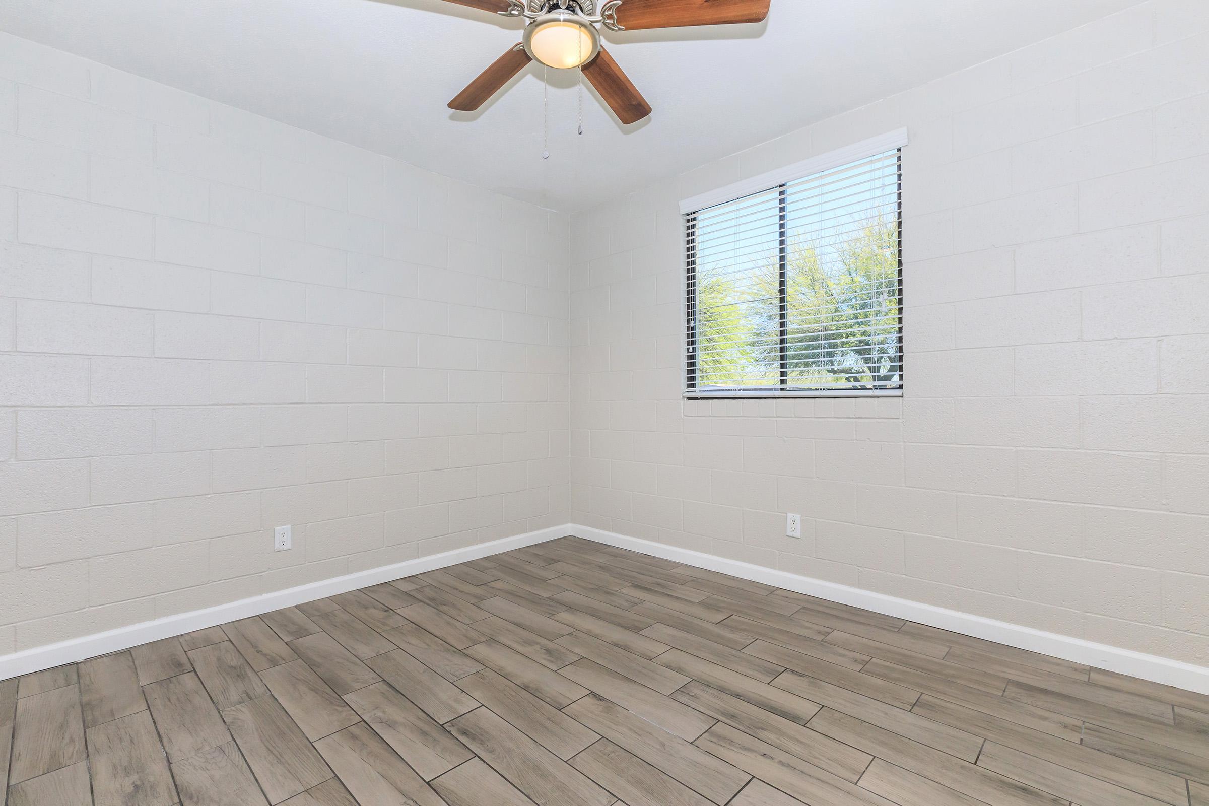 Empty room with light-colored walls and a tiled floor. A ceiling fan with wooden blades is installed. The room features a window with horizontal blinds, allowing natural light to enter, and is sparsely furnished, creating a minimalistic feel.