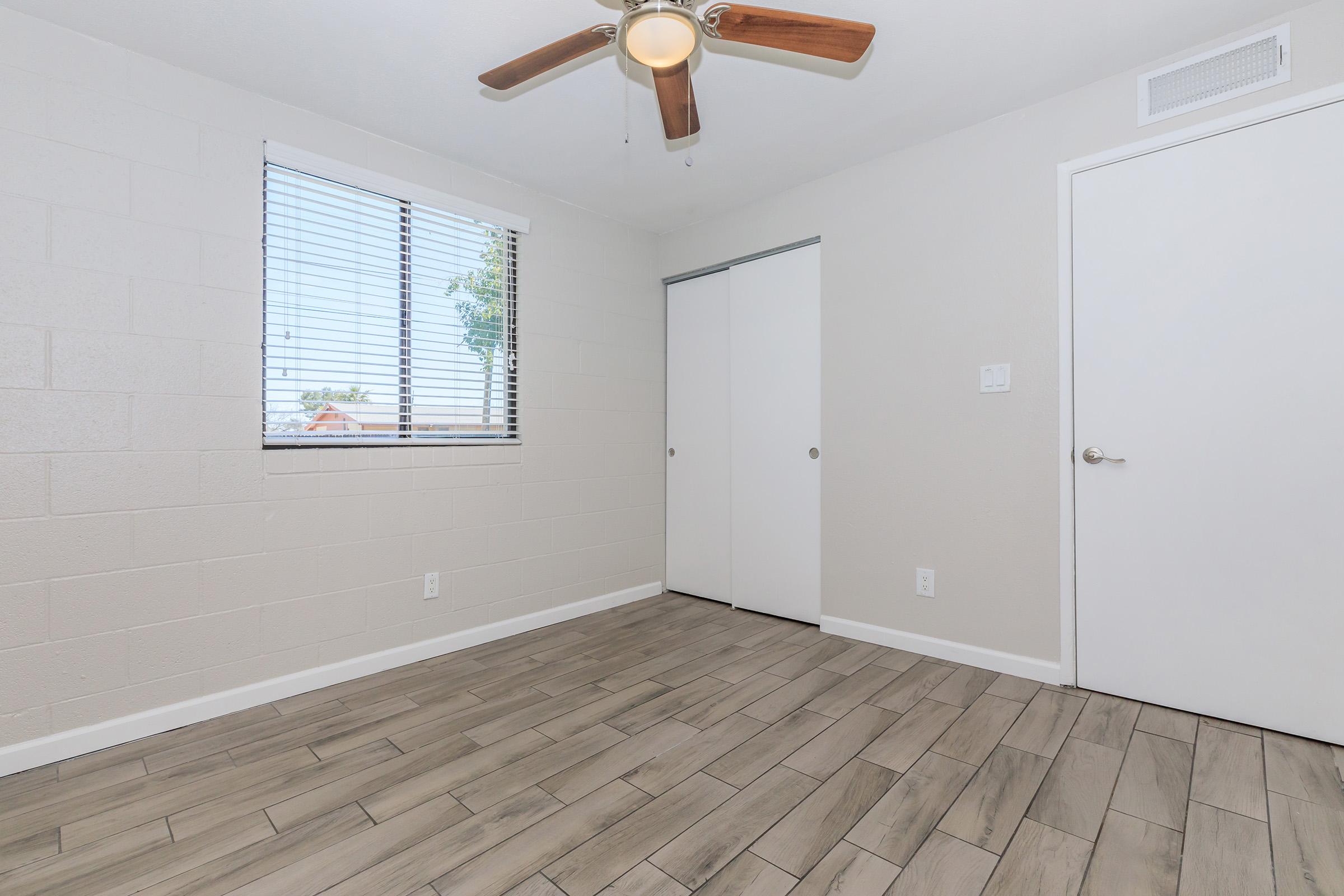 A clean, minimalistic room featuring light-colored walls and a wooden floor. The space includes a ceiling fan with wooden blades, a window with blinds allowing natural light, and a closet with sliding doors. A simple white door is visible on the right side.