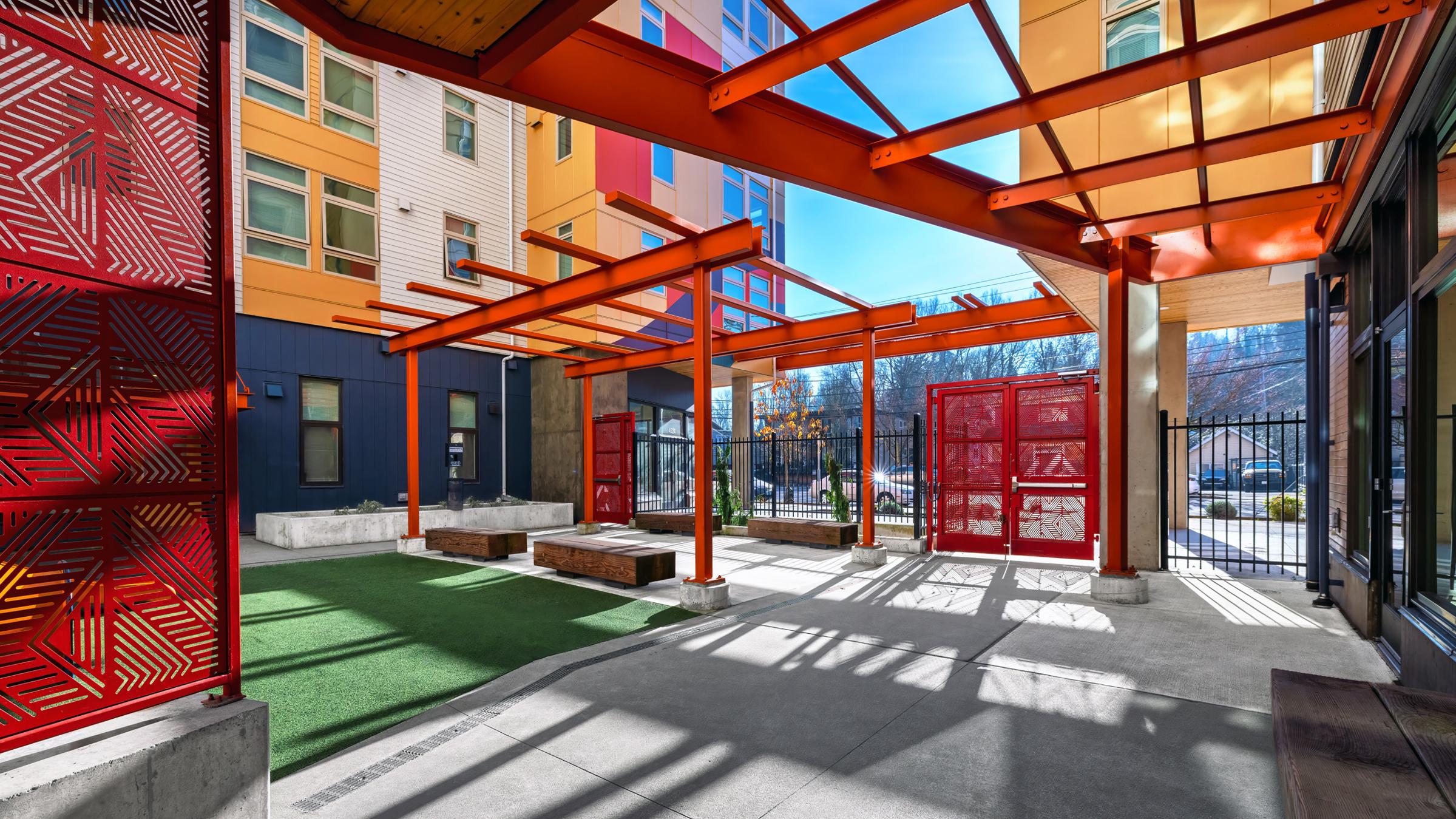 A modern outdoor space featuring colorful, geometric structures with red metal accents and transparent roofs. The area includes green grass surfaces, concrete pathways, and is surrounded by contemporary apartment buildings. Sunlight casts playful shadows across the space, creating a bright and inviting atmosphere.