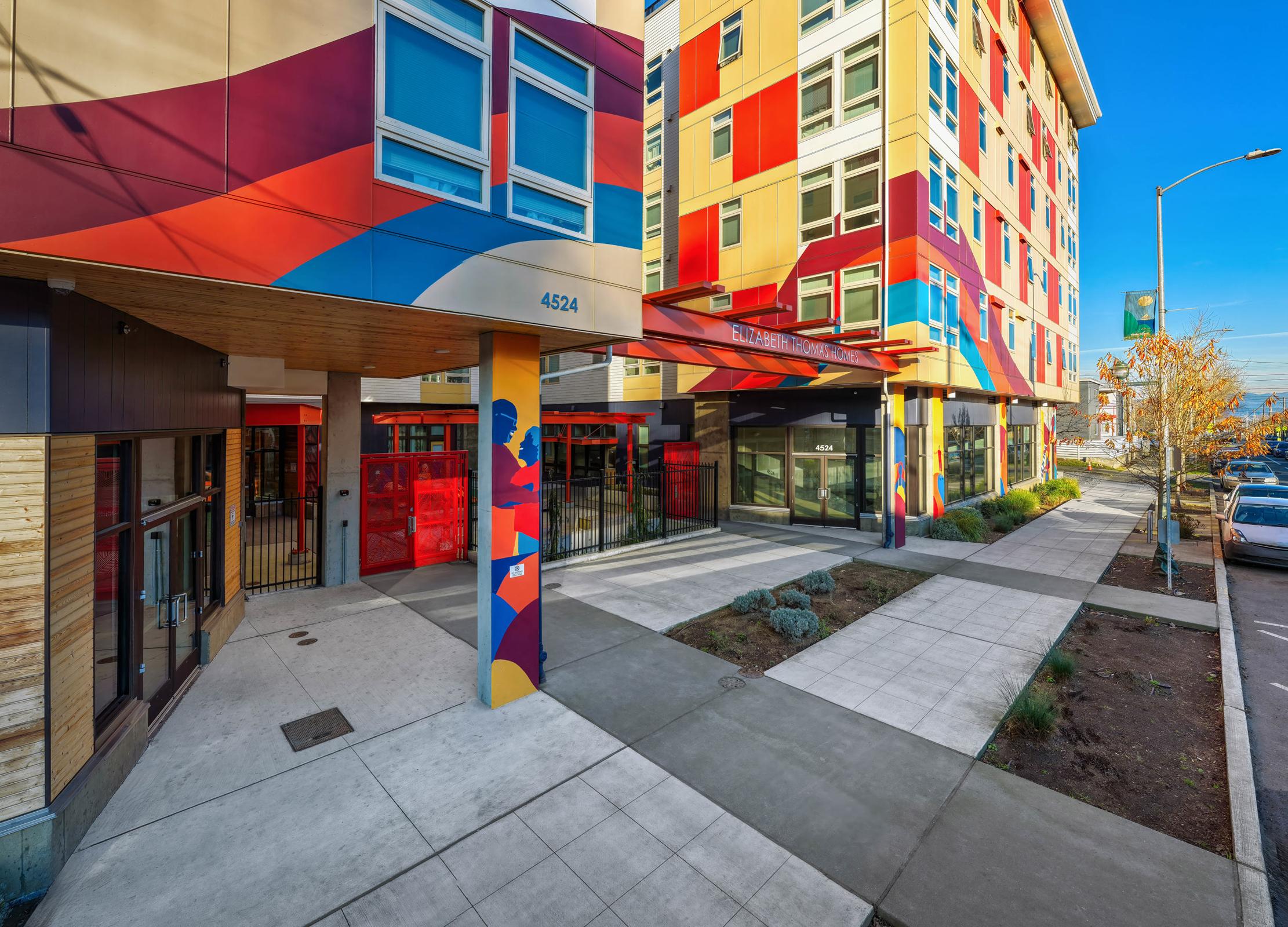 Vibrant modern building exterior featuring colorful patterns on the facade, with a welcoming entrance marked by bold artwork. The area includes landscaped pathways and a tree-lined street, contributing to an inviting urban environment. Clear blue sky overhead.