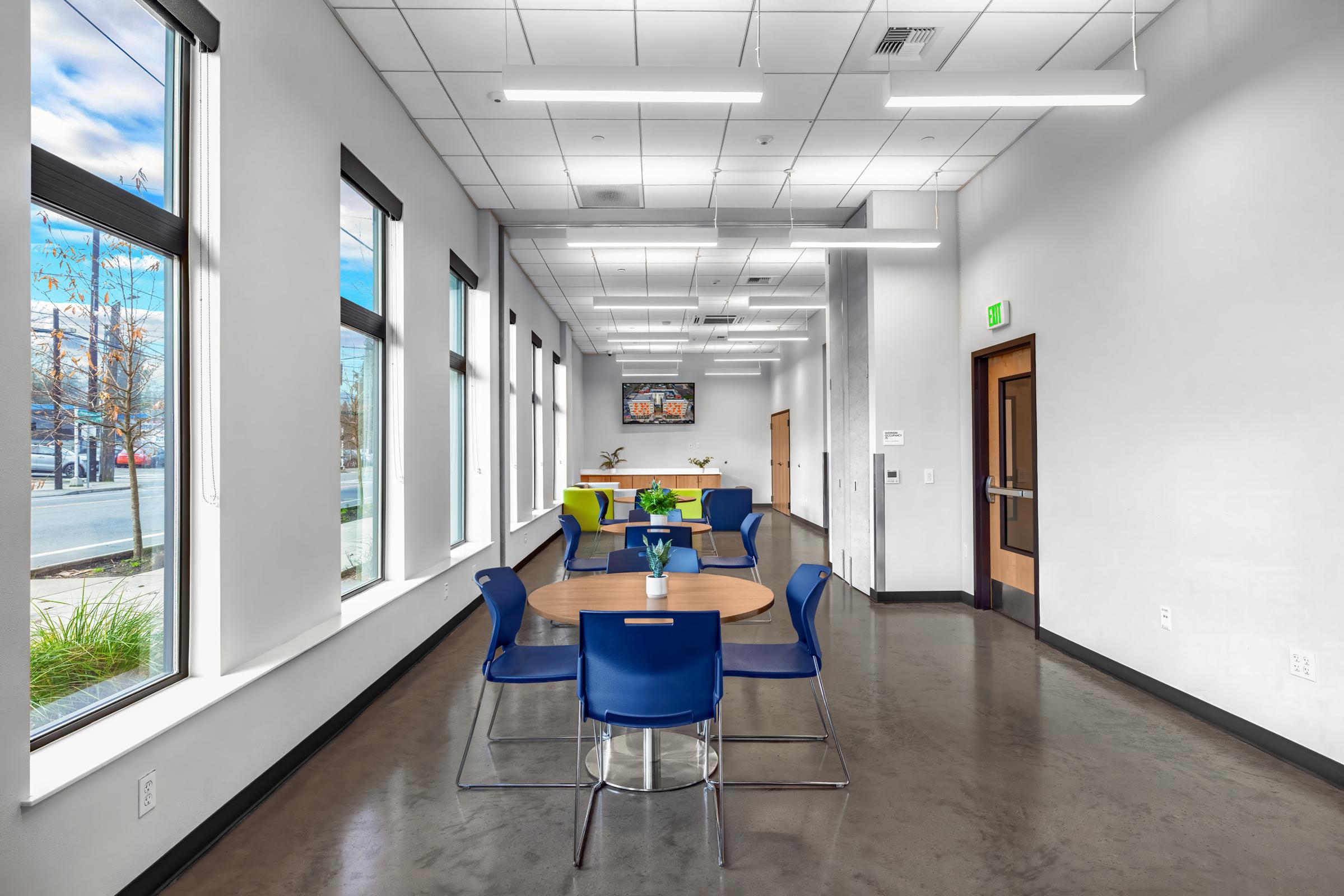 A modern interior space featuring a long hallway with large windows, allowing natural light to flood in. On one side, there is a round table with blue chairs arranged around it, and a decorative plant centerpiece. At the far end, there is a TV mounted on the wall and bright green seating area. The floor is polished concrete.