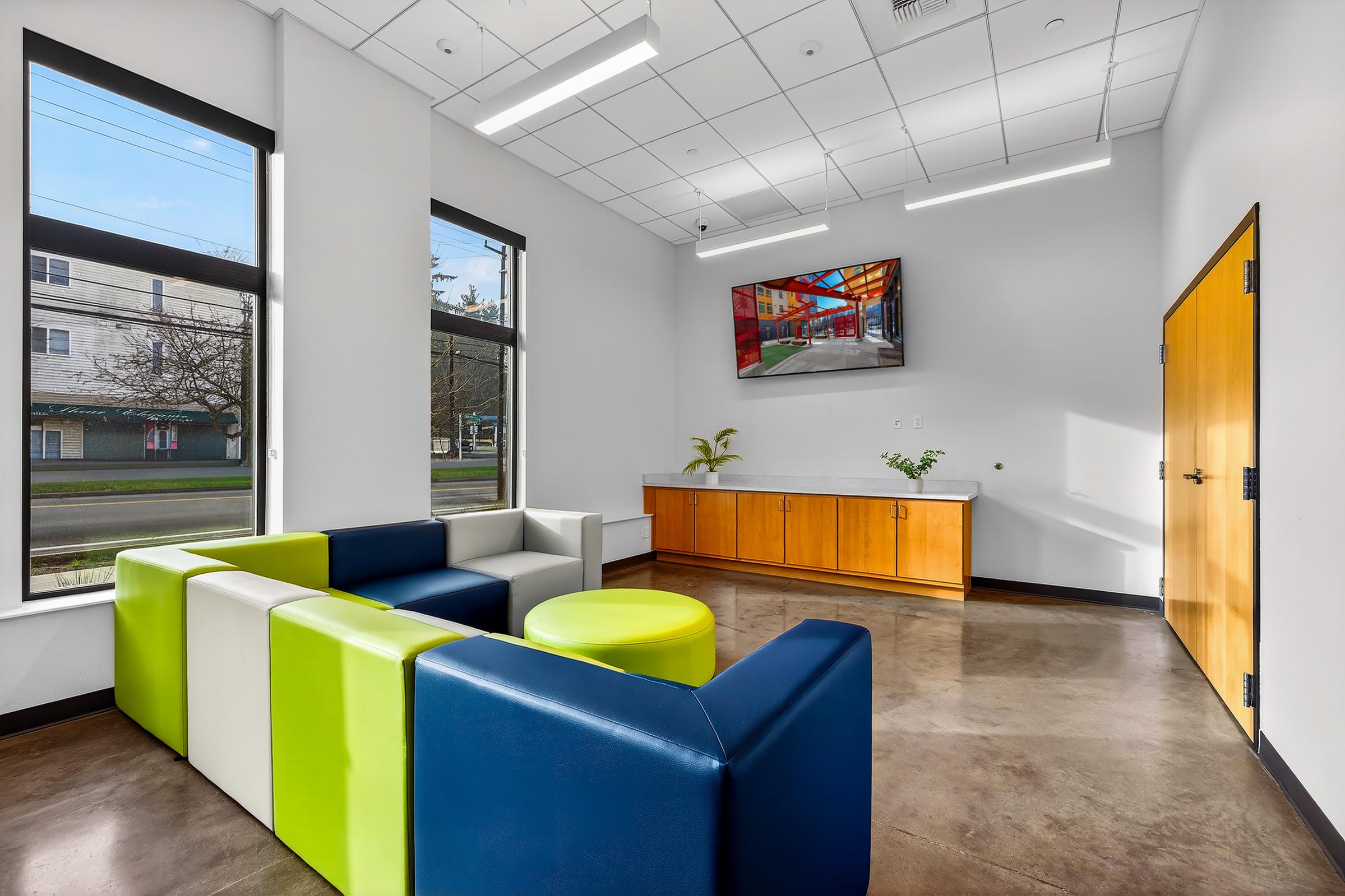 A modern lounge area featuring a multi-colored sectional couch in blue, green, and white, a round coffee table, and a wooden cabinet against the wall. Large windows provide natural light, and a mounted TV displays colorful artwork. The floor is polished concrete, creating a sleek, contemporary vibe.