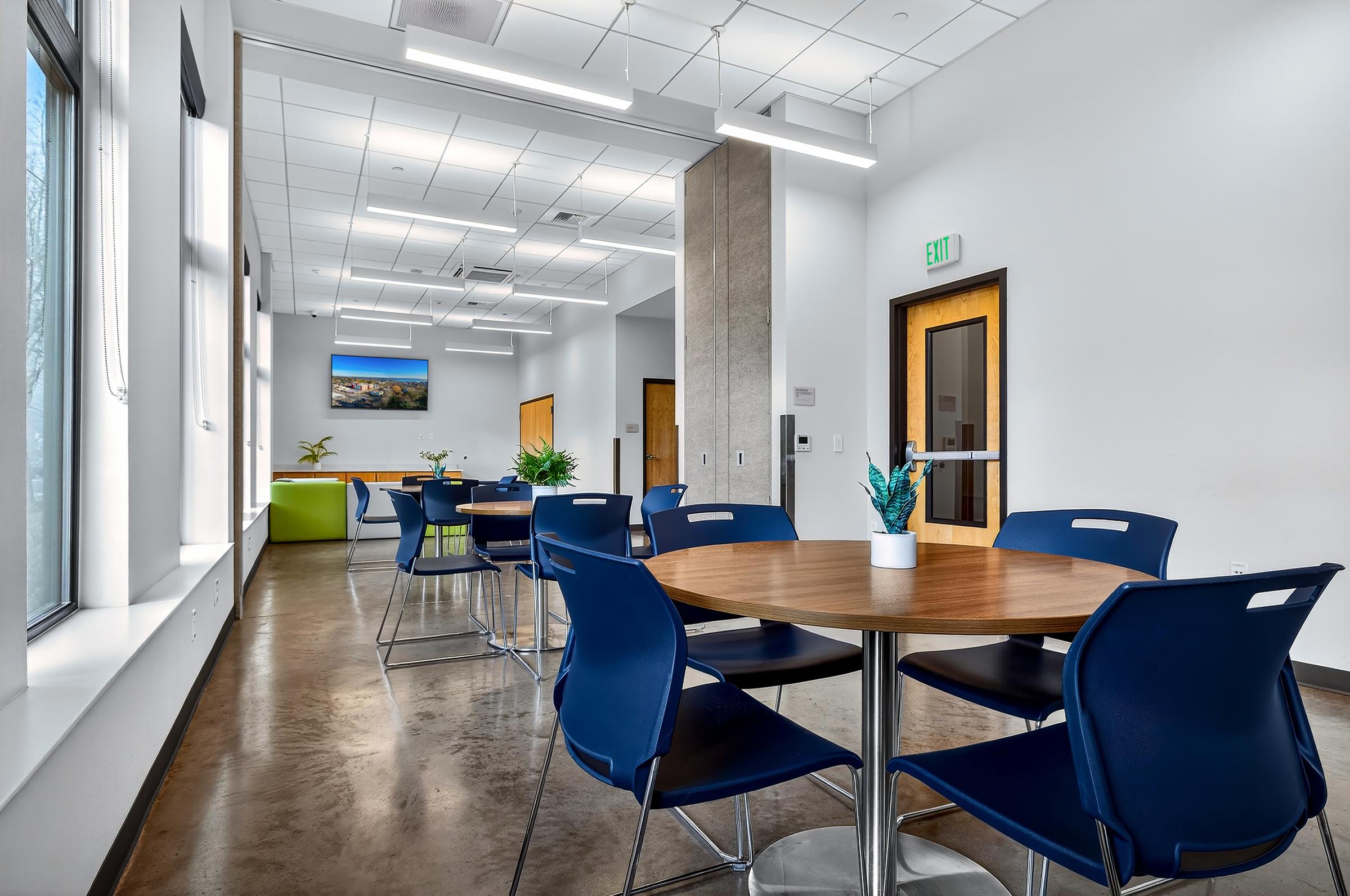 A modern, well-lit interior of a communal space featuring several tables and chairs. The design includes a round table with a small potted plant, blue chairs, and large windows allowing natural light. In the background, there is a landscape painting on the wall and a green couch.