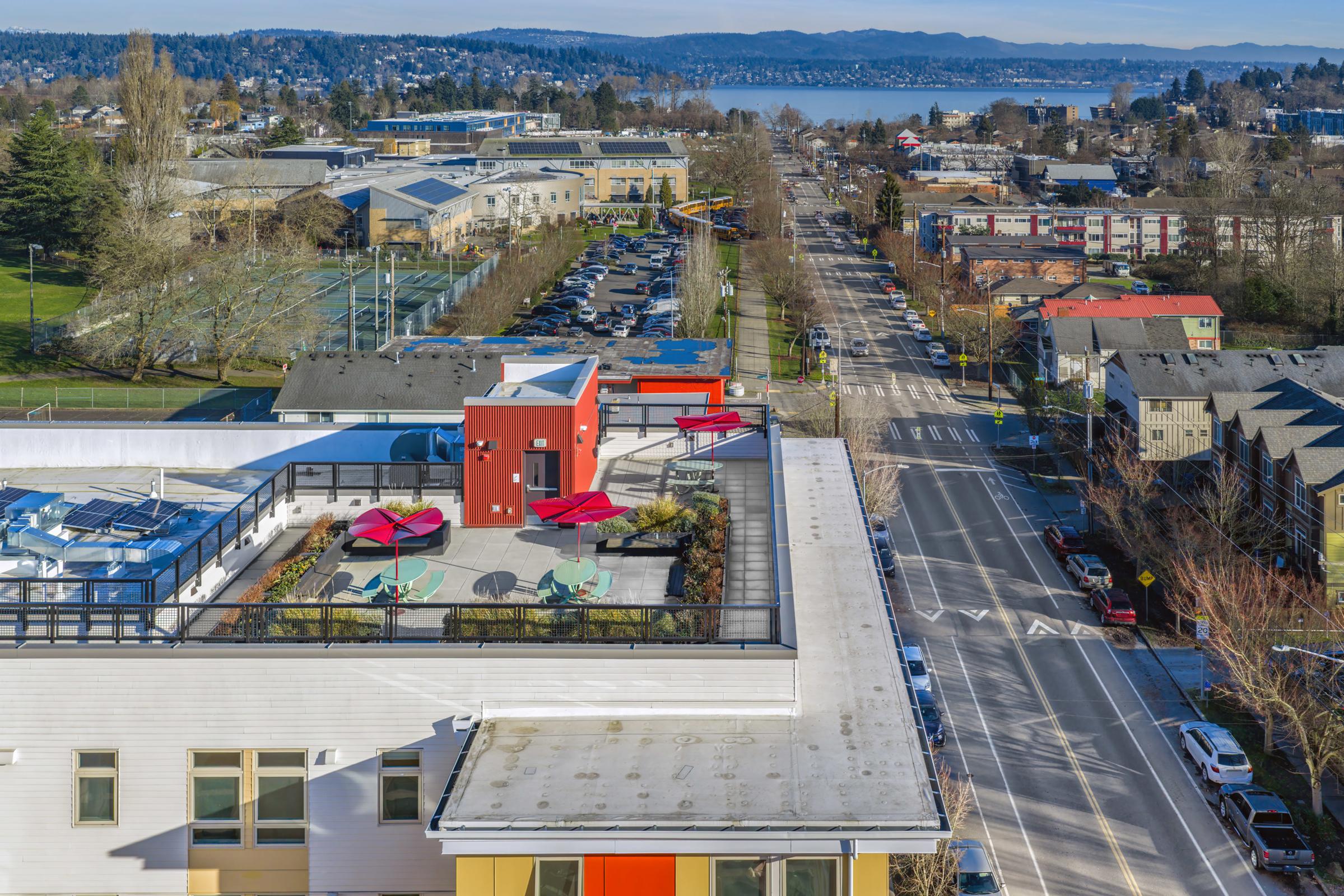 Aerial view of a neighborhood featuring a rooftop terrace with red umbrellas and green chairs. In the background, there are residential buildings and a scenic waterfront with mountains in the distance. Streets are lined with parked cars and trees, showcasing an urban landscape on a clear day.
