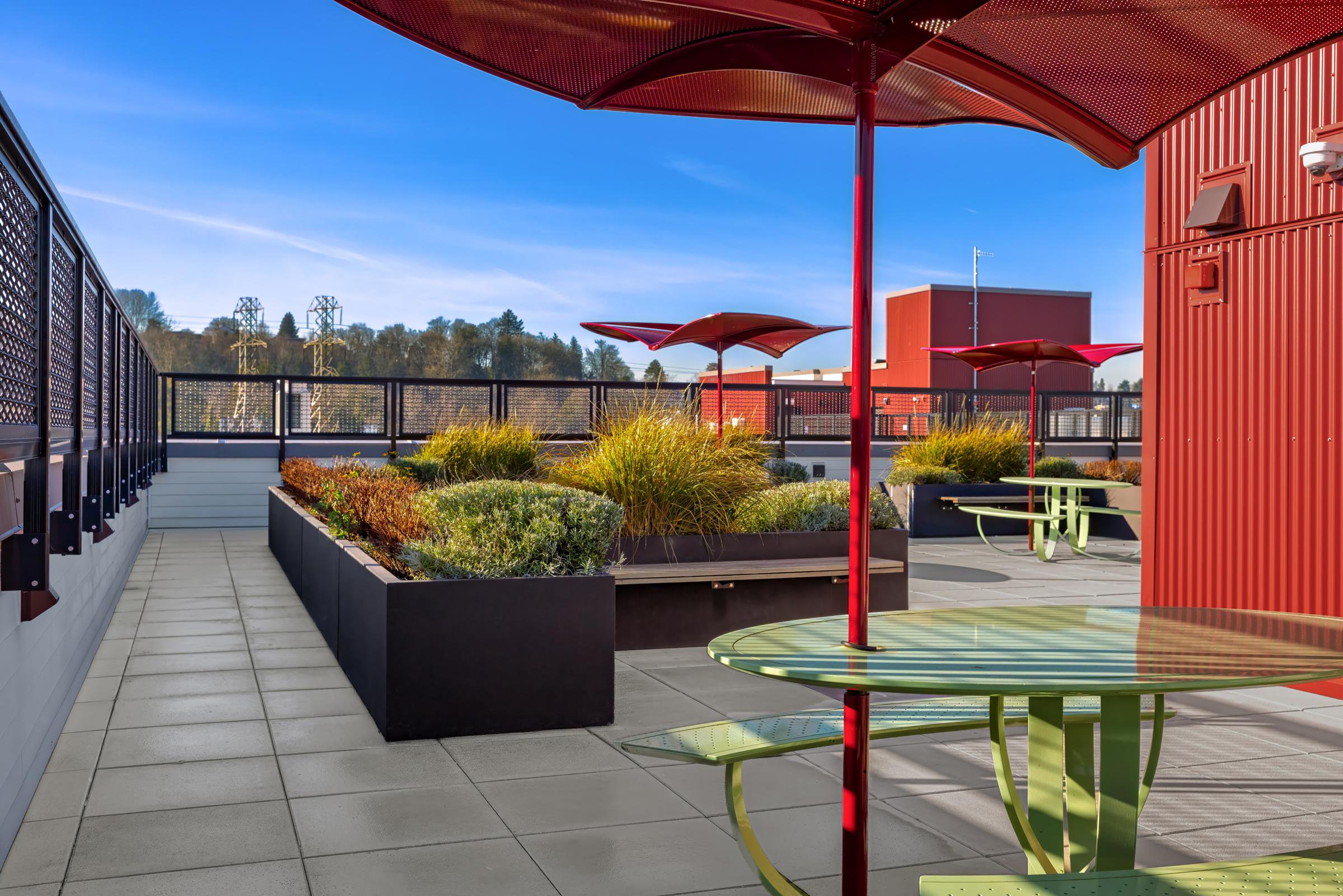 A modern rooftop terrace featuring vibrant red parasols, green tables, and raised garden beds filled with ornamental grasses. The space is designed for relaxation, with a clear blue sky and a railing offering views of the surrounding area. The contemporary architecture includes red paneling and a stylish layout.