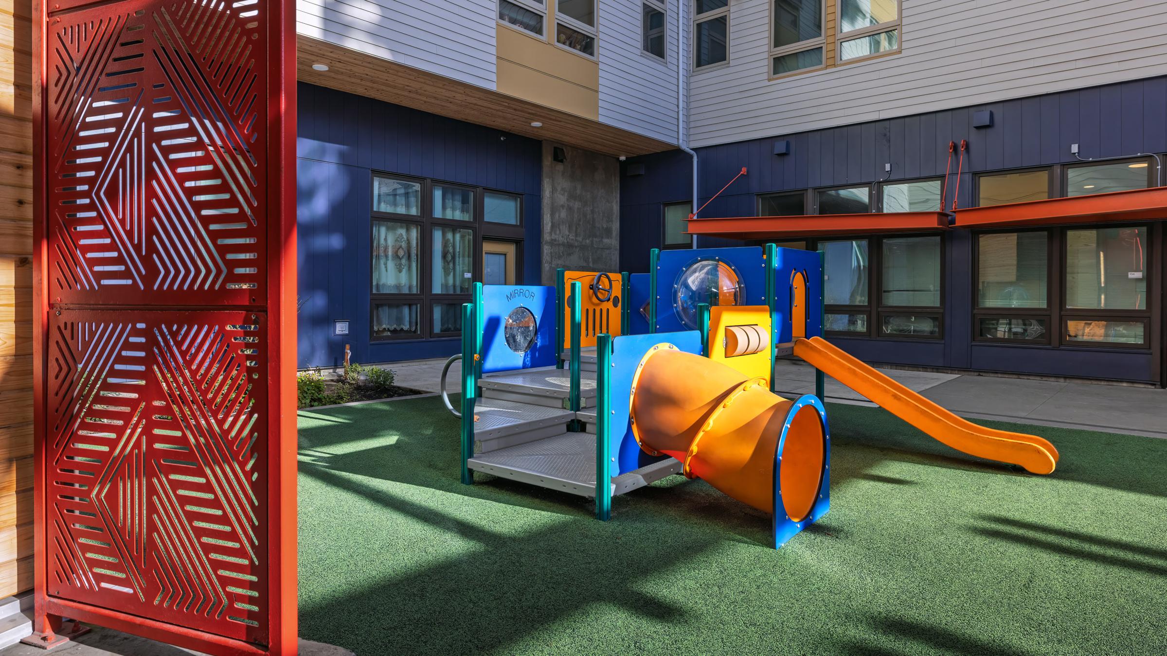 Colorful playground equipment featuring a slide, a tunnel, and climbing structures, situated on a grassy area. The surrounding building has large windows and modern design elements, with a decorative red panel visible on the left. The scene is bright and inviting, ideal for children's play.