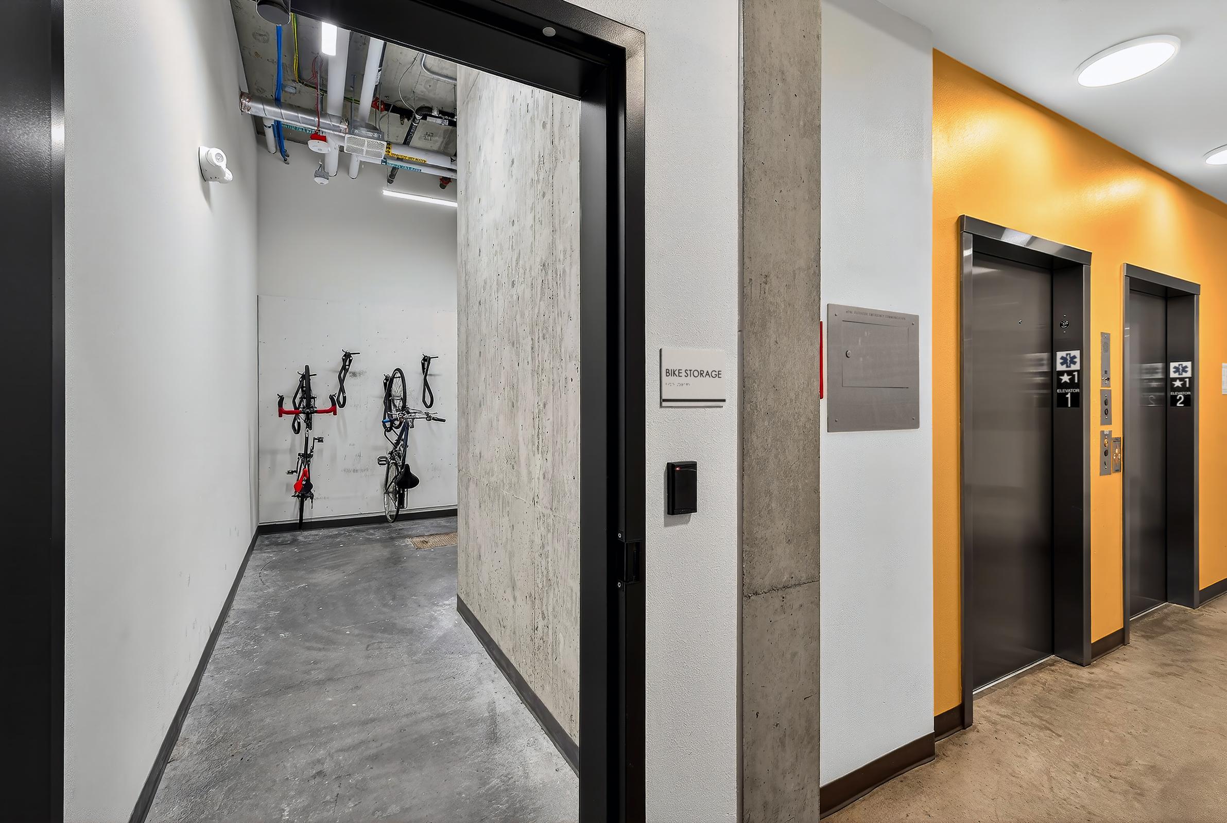 A hallway featuring bike storage on the left with bike racks mounted on the wall, and two elevator doors on the right. The walls are a mix of concrete and a bright yellow, with overhead lighting creating a modern atmosphere.