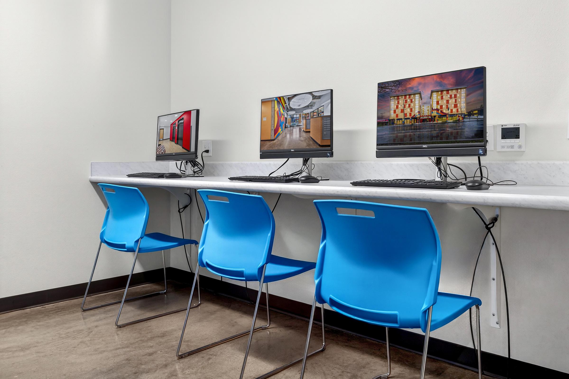 Three desktop computers with monitors are positioned on a long white countertop. Each workstation is accompanied by a blue chair. The background features a plain white wall and a visible cable management system.