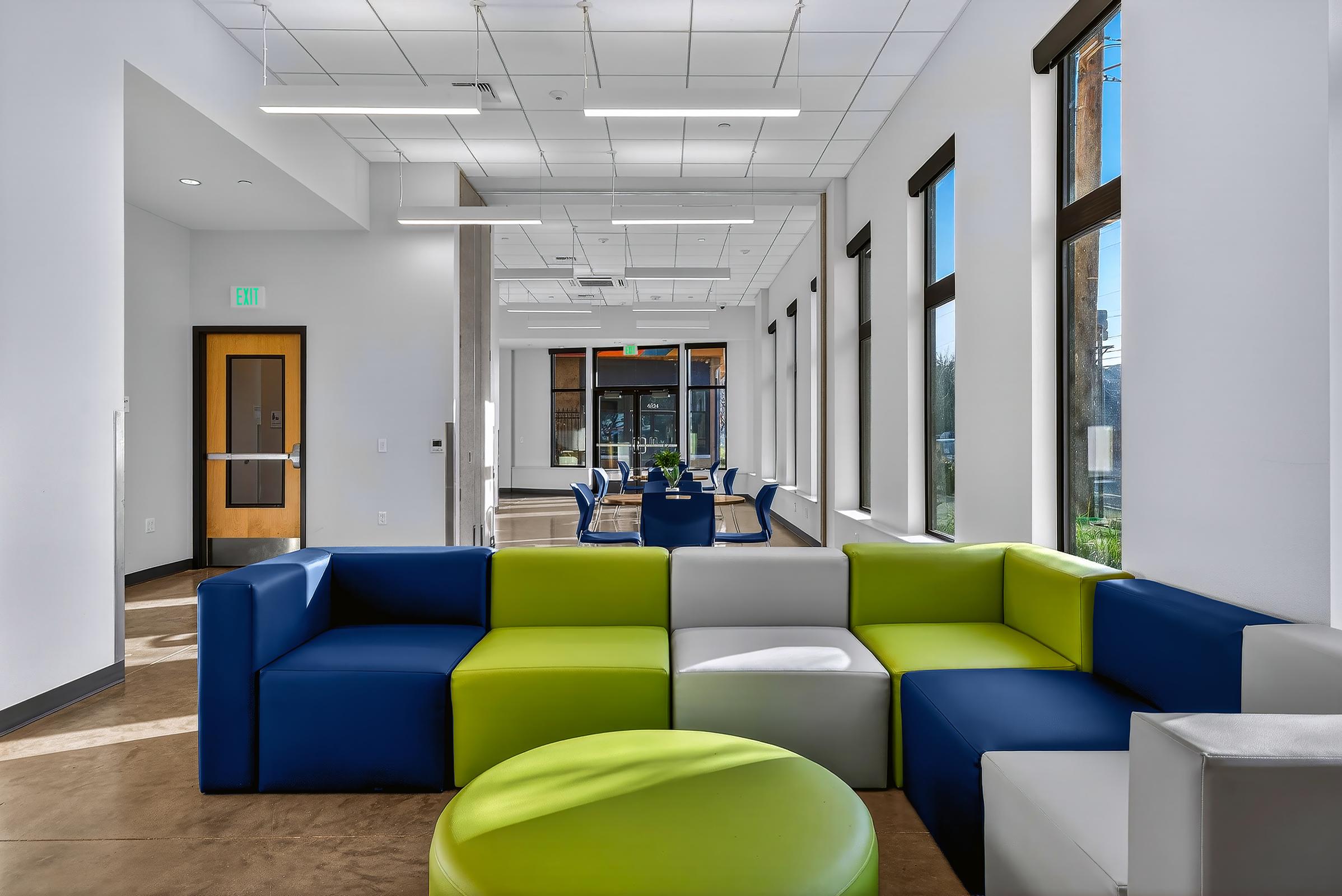 Modern interior of a spacious lounge area featuring colorful furniture, including a blue, green, and white sectional couch and round green ottomans. Large windows allow natural light to brighten the space, which has contemporary lighting and a clean, minimalist design.