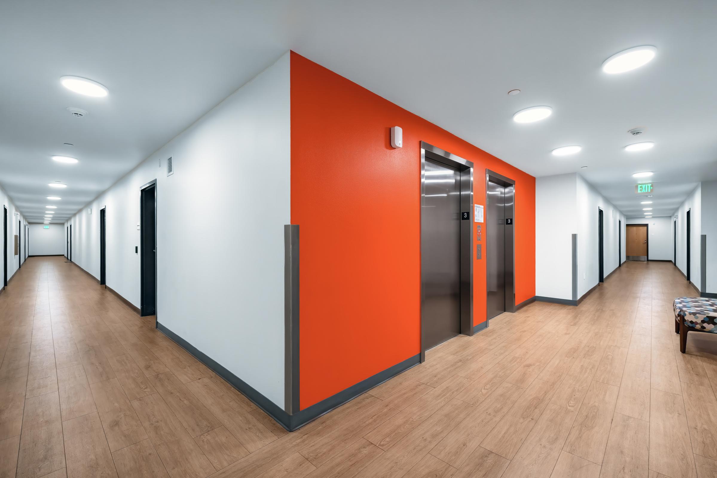 Modern interior hallway in a building featuring a vibrant orange accent wall, two elevators at the end, polished wooden floors, and doors leading to various rooms on both sides. Soft lighting creates a welcoming atmosphere, with a small seating area visible in the foreground.