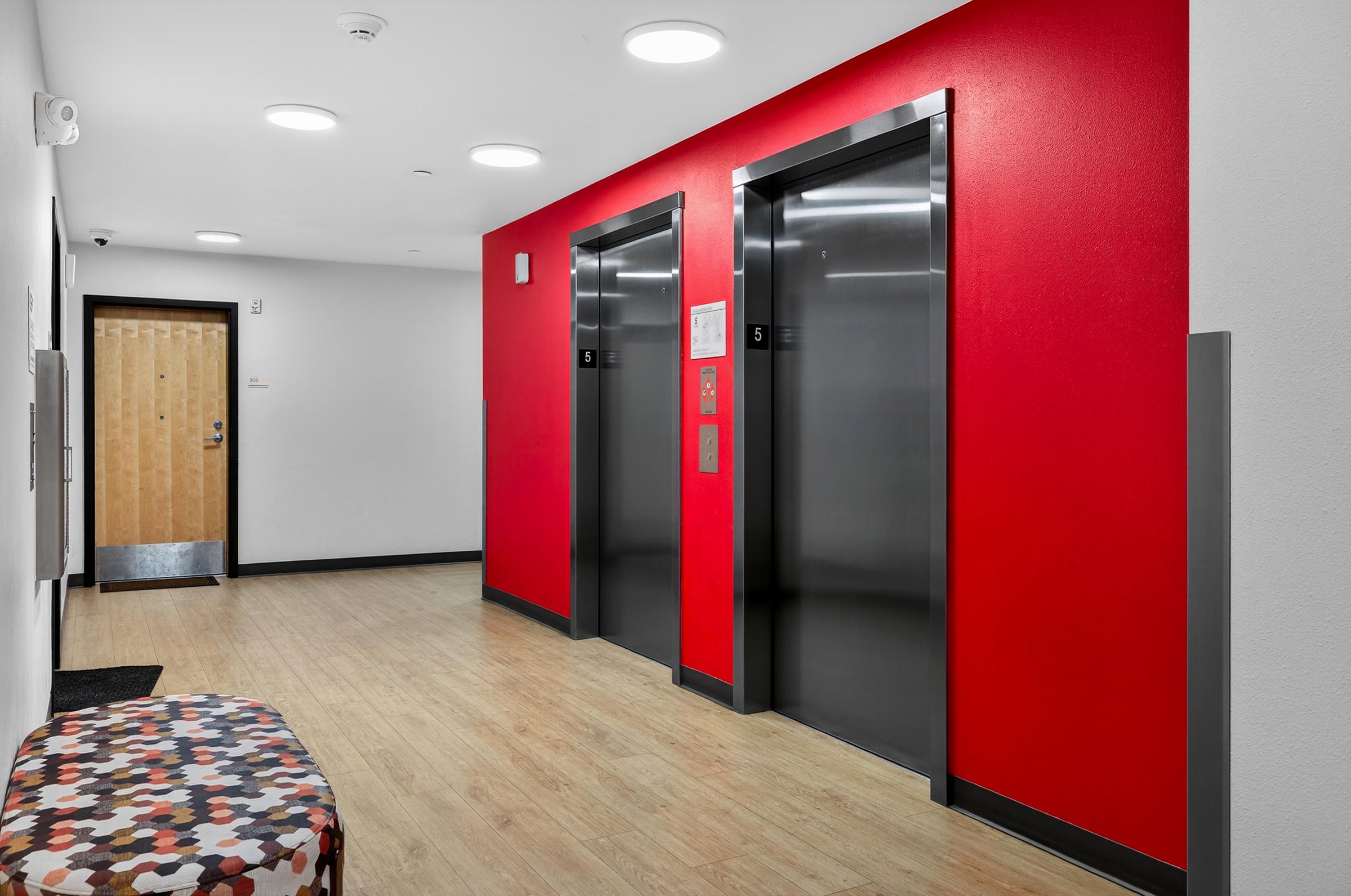 A modern hallway featuring two elevators with sleek metal doors against a bright red wall. A colorful, patterned bench is positioned to the side on a light wood floor. A wooden door is visible at the end of the hallway, along with a wall-mounted mailbox or directory.