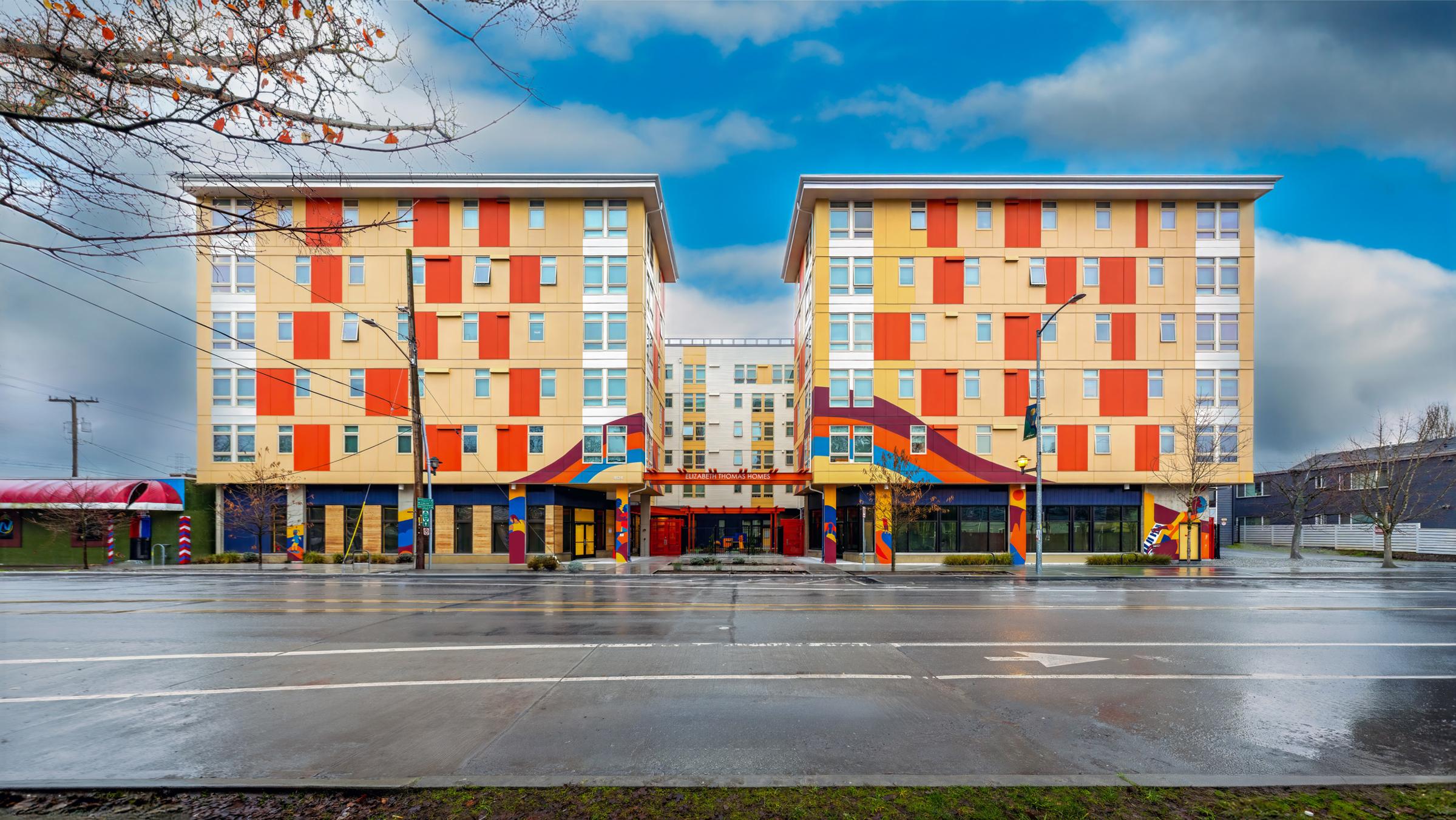 Vibrant modern apartment complex with colorful orange, yellow, and blue façade. The building features large windows and a welcoming entrance, set against a cloudy sky. The street in front is empty and wet, suggesting recent rain. The architecture combines contemporary design with bold colors.