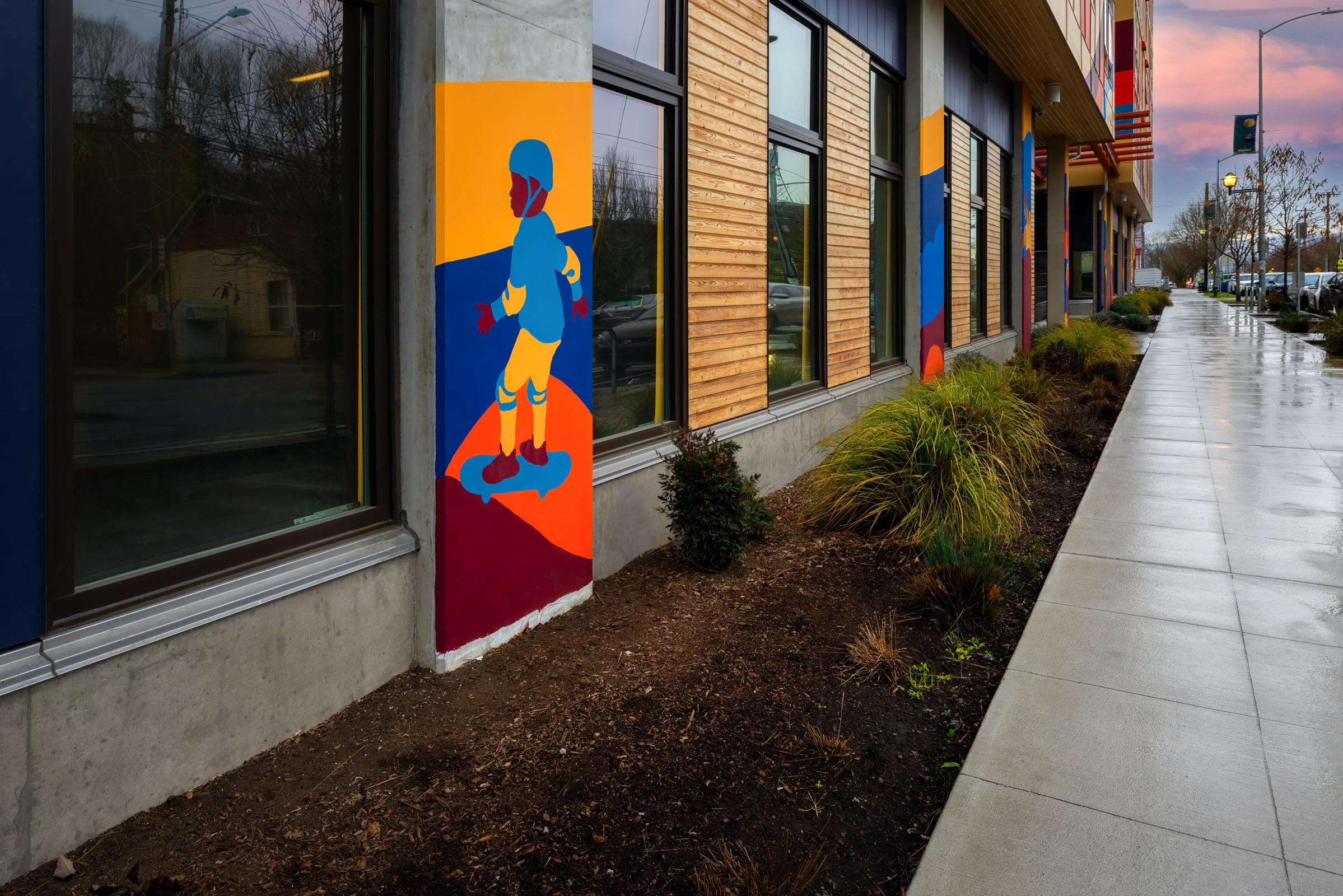 Colorful street art depicting a cartoon figure on a skateboard, positioned on the exterior walls of a modern building. The artwork features vibrant colors, contrasting with the natural surroundings, and the sidewalk is wet, reflecting the overcast sky. Lush greenery lines the path.